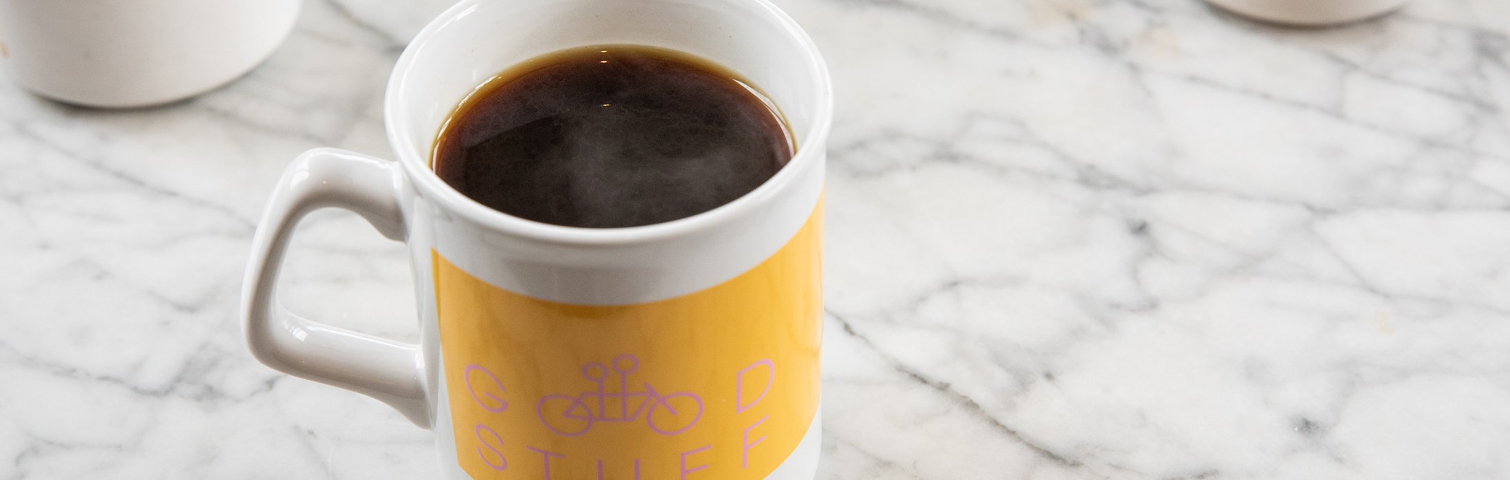 A yellow and white coffee mug with the words "Good Stuff" partially visible, filled with steaming black coffee, placed on a white marble countertop.