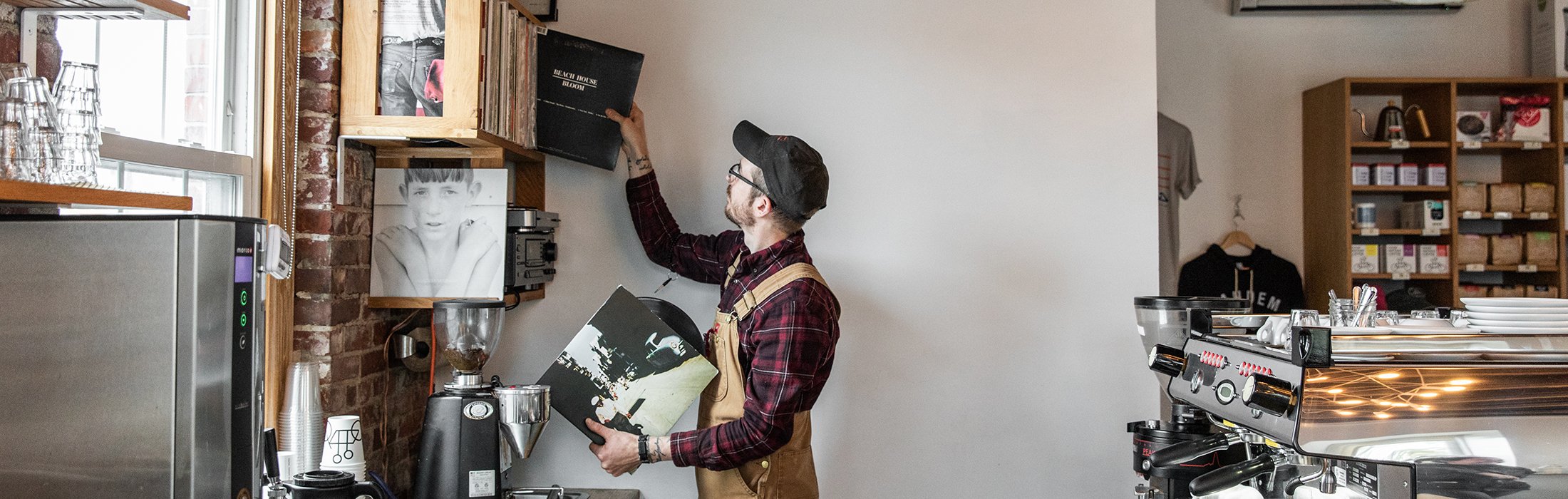A barista in a coffee shop wearing an apron and cap reaches up to place a vinyl record on a shelf. The shelves contain other records and coffee equipment. A coffee machine is seen on the right, and window light brightens the scene.