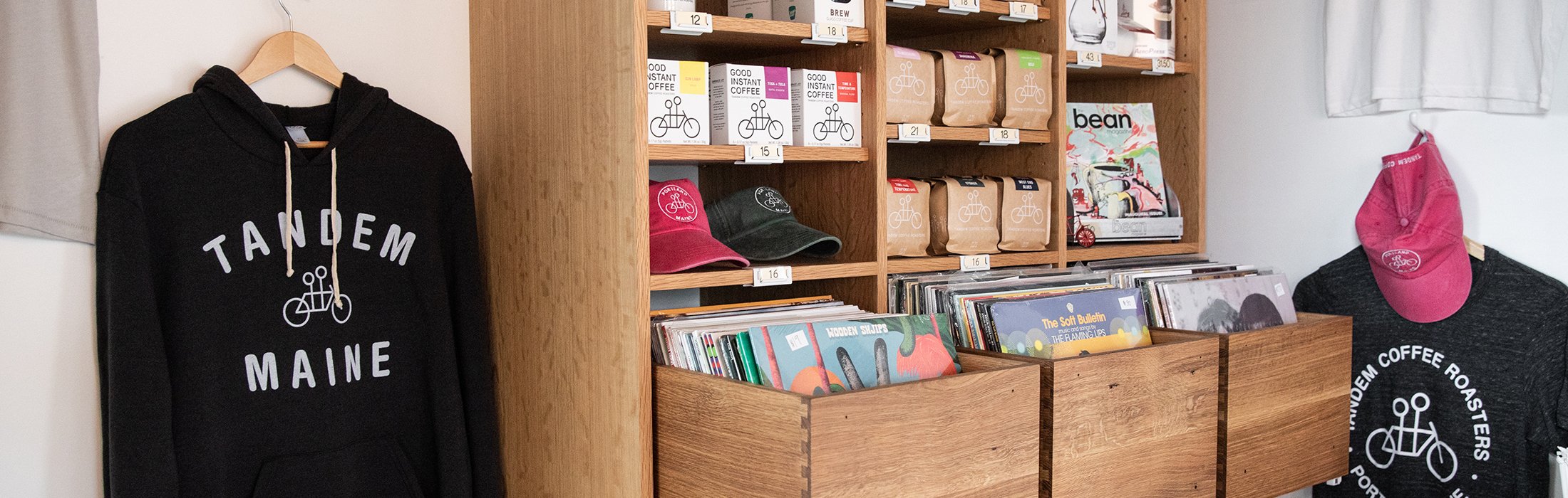 A display shelf with coffee bags, branded caps, T-shirts, and vinyl records. A black hoodie with "Tandem Maine" hangs on the wall. Shelves are neatly organized.