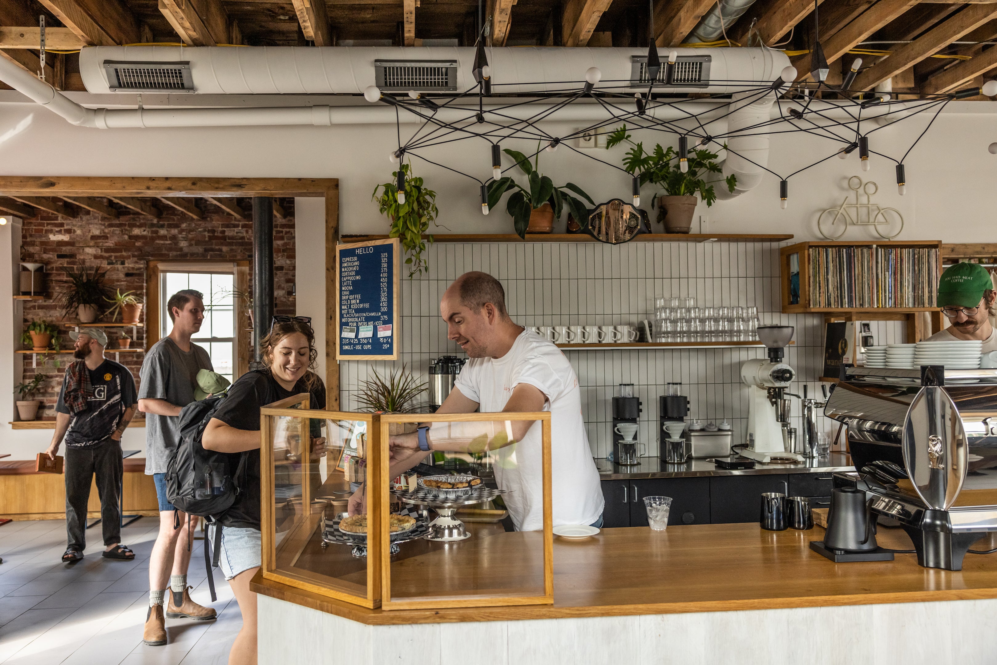 A cozy café interior with customers standing at the counter where a barista is serving, surrounded by rustic brick walls, wooden beams, and plants hanging above.