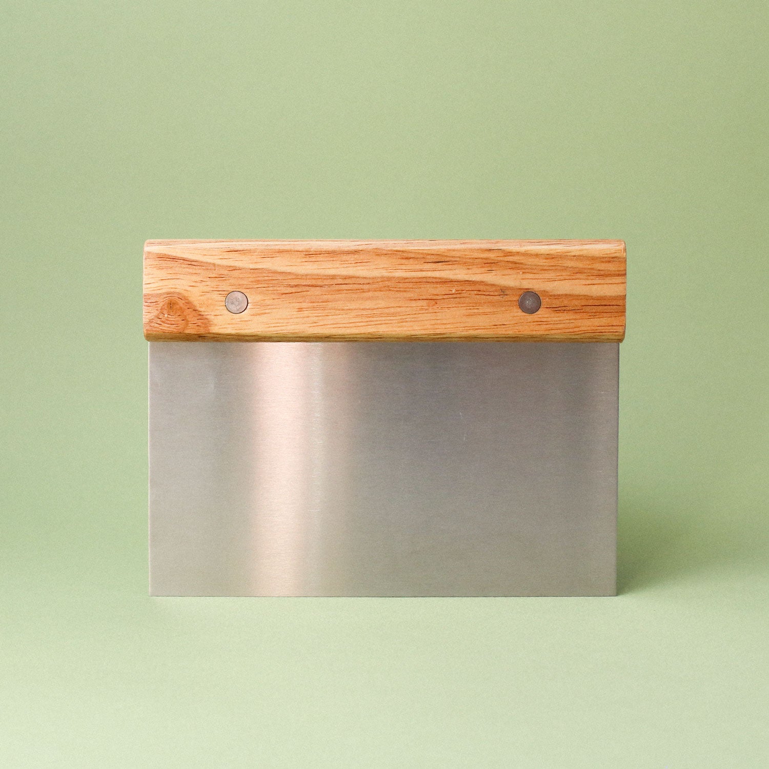 The Tandem Bench Scraper by Tandem Coffee Roasters, featuring a metal blade and wooden handle, stands upright on a light green surface against a matching green background—ideal for dough handling.
