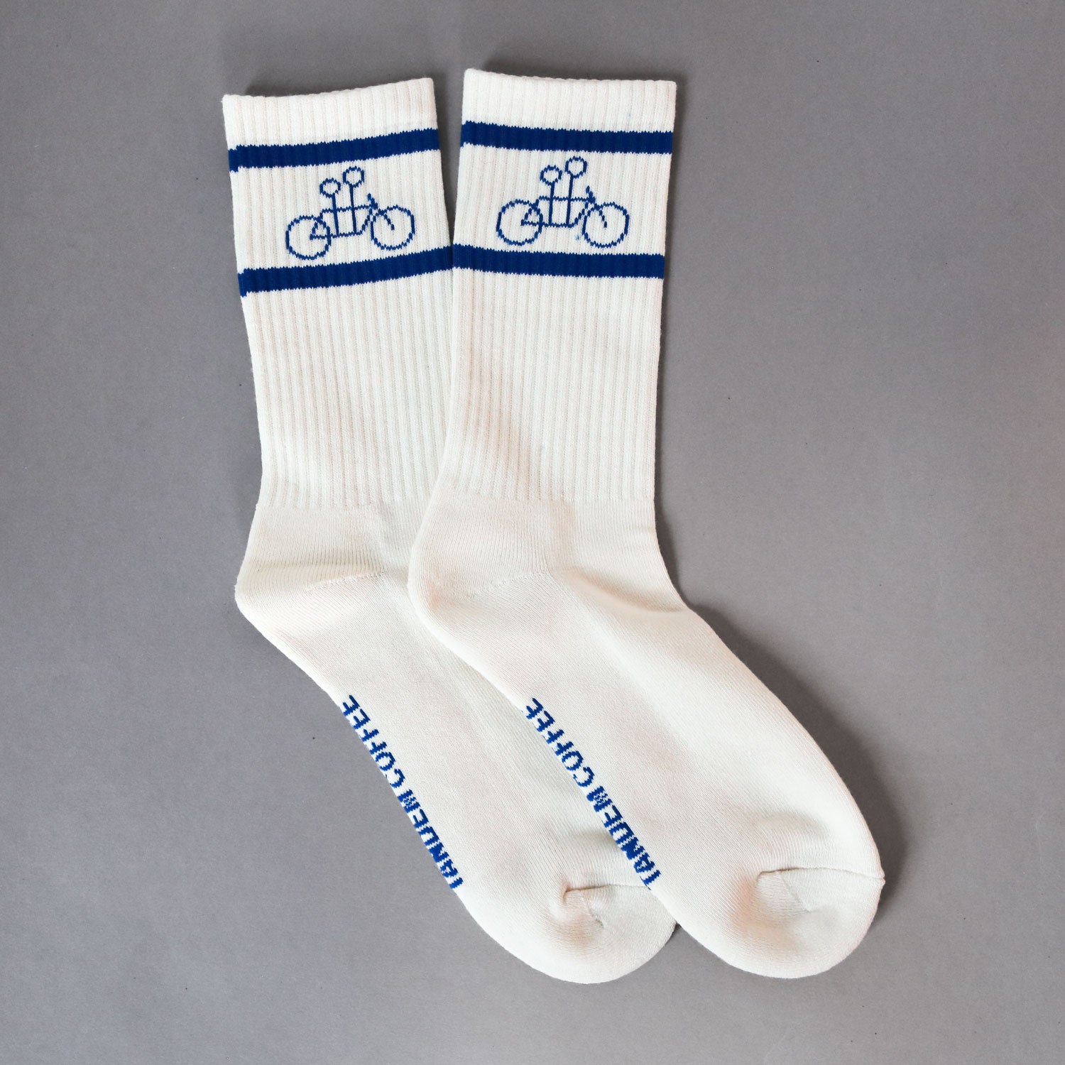 The Tandem Socks from Tandem Coffee Roasters feature a simple blue bicycle design with dark blue stripes near the top on white cotton. Elegantly displaying "Tandem Coffee" across the toes, these socks are laid flat on a gray background, seamlessly blending style and comfort.