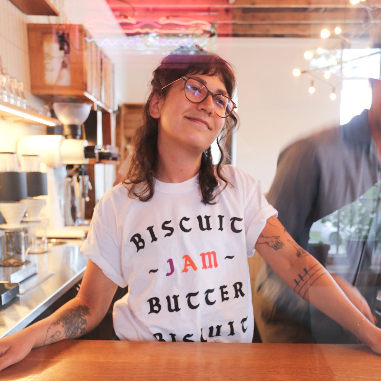 A young woman with glasses, wearing a Tandem Coffee Roasters The Loaded Biscuit Tee, stands confidently in a cozy room with wooden shelves filled with boxes behind her.