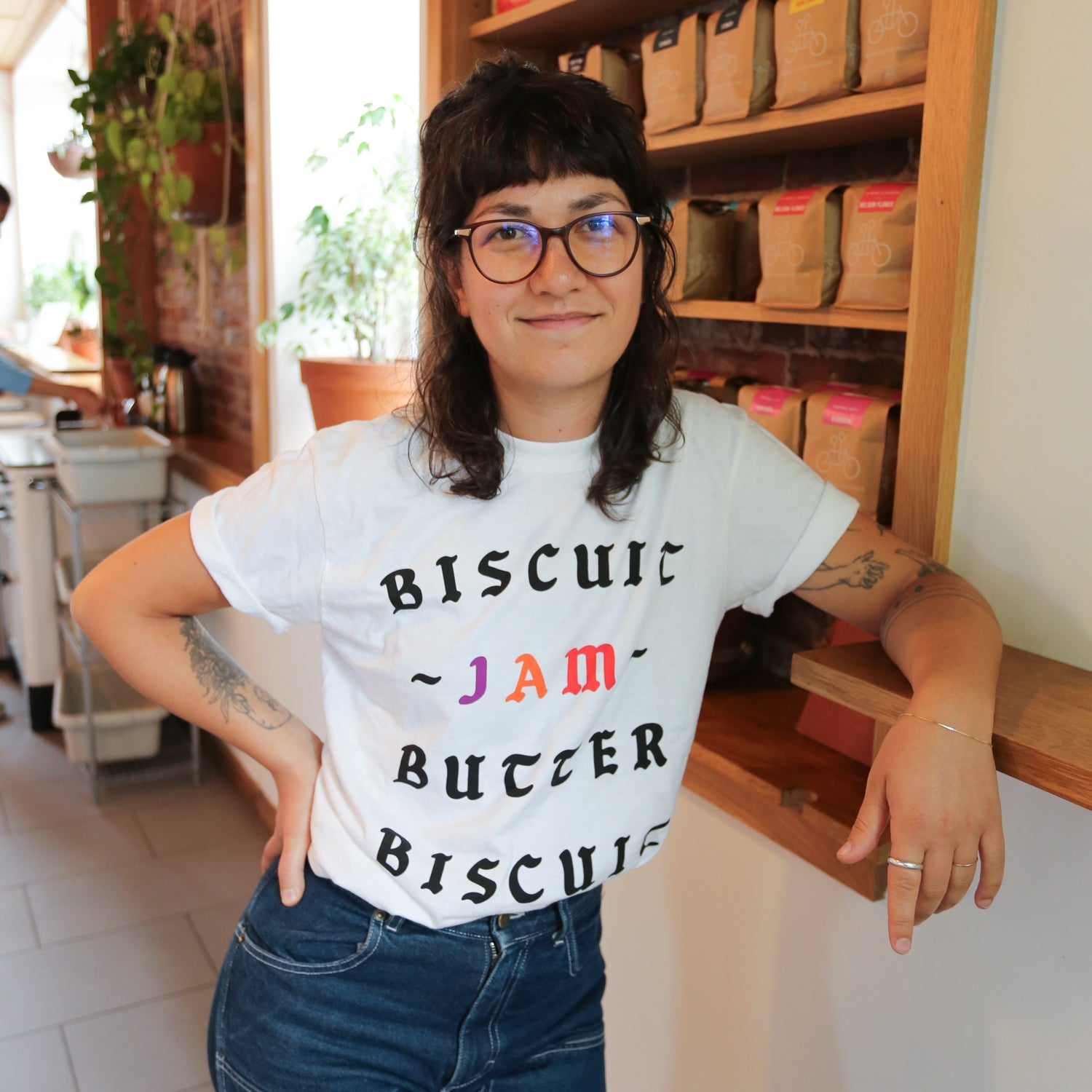 A young woman with glasses, wearing a Tandem Coffee Roasters The Loaded Biscuit Tee, stands confidently in a cozy room with wooden shelves filled with boxes behind her.