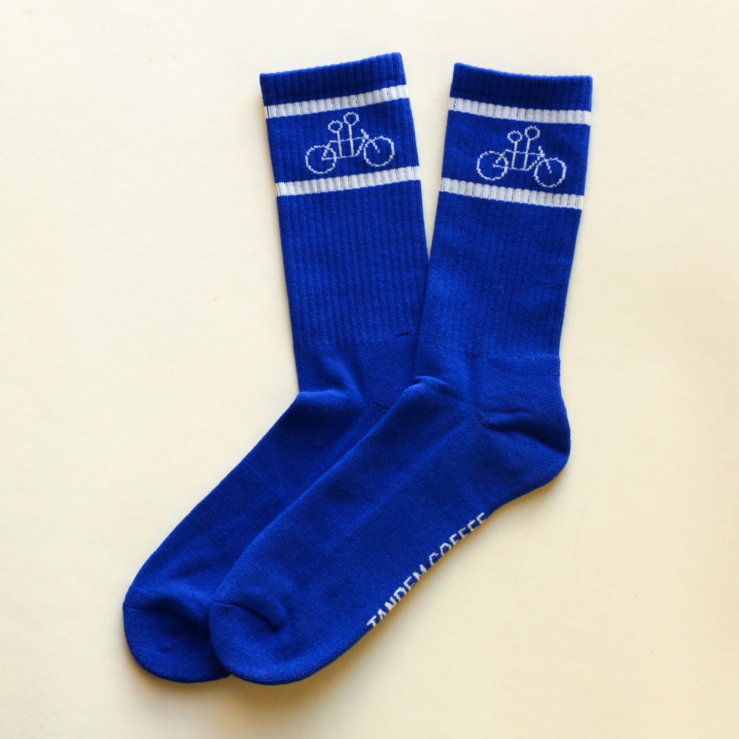The Tandem Socks by Tandem Coffee Roasters are one size, blue with white stripes, feature a tandem bike graphic, and have "TANDEM COFFEE" printed in white on the sole of one sock.