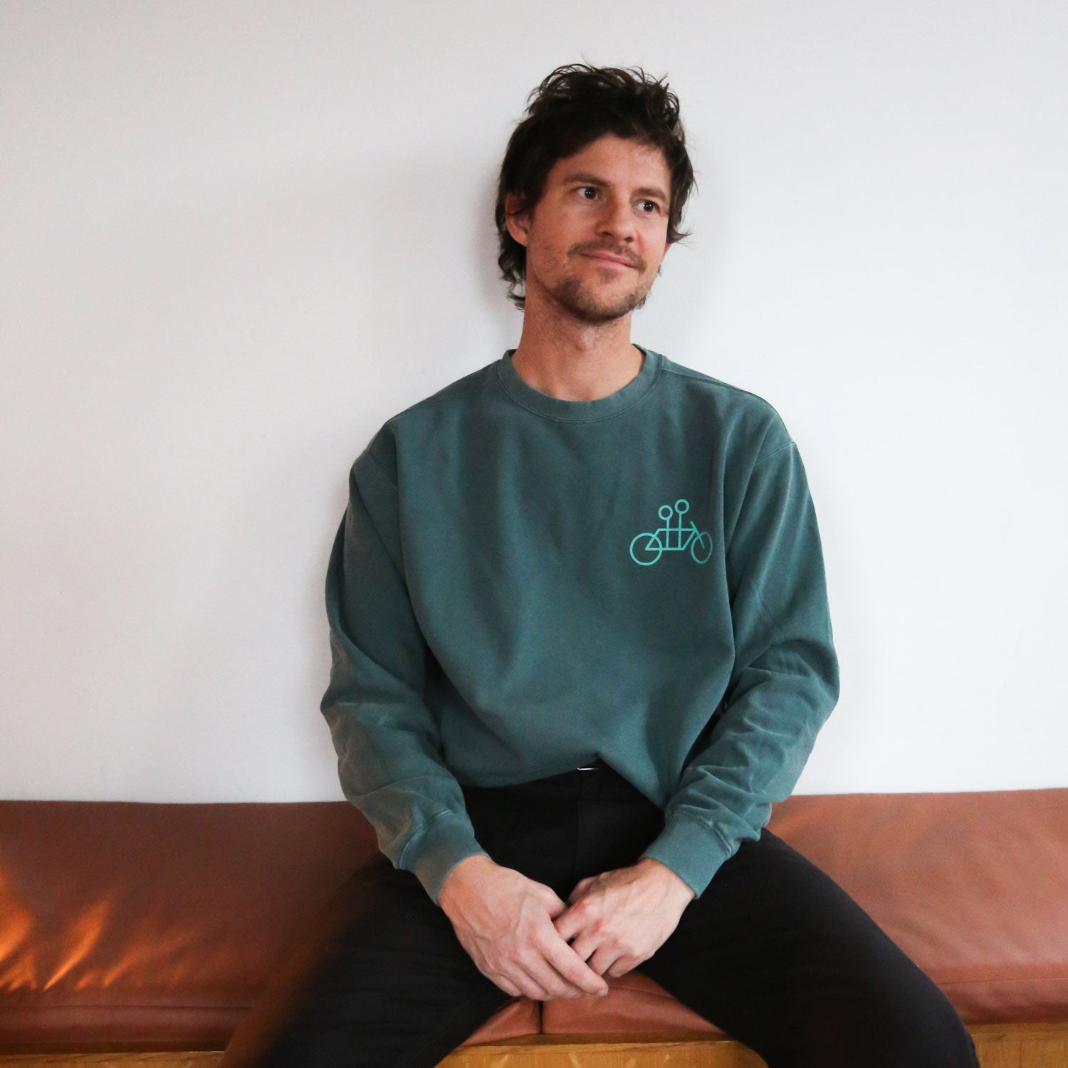 A person with short, dark hair is wearing a cozy Blue Spruce Tandem Crewneck from Tandem Coffee Roasters, featuring a bicycle graphic. They are sitting on a brown leather bench against a plain white wall, smiling and looking slightly to the side.