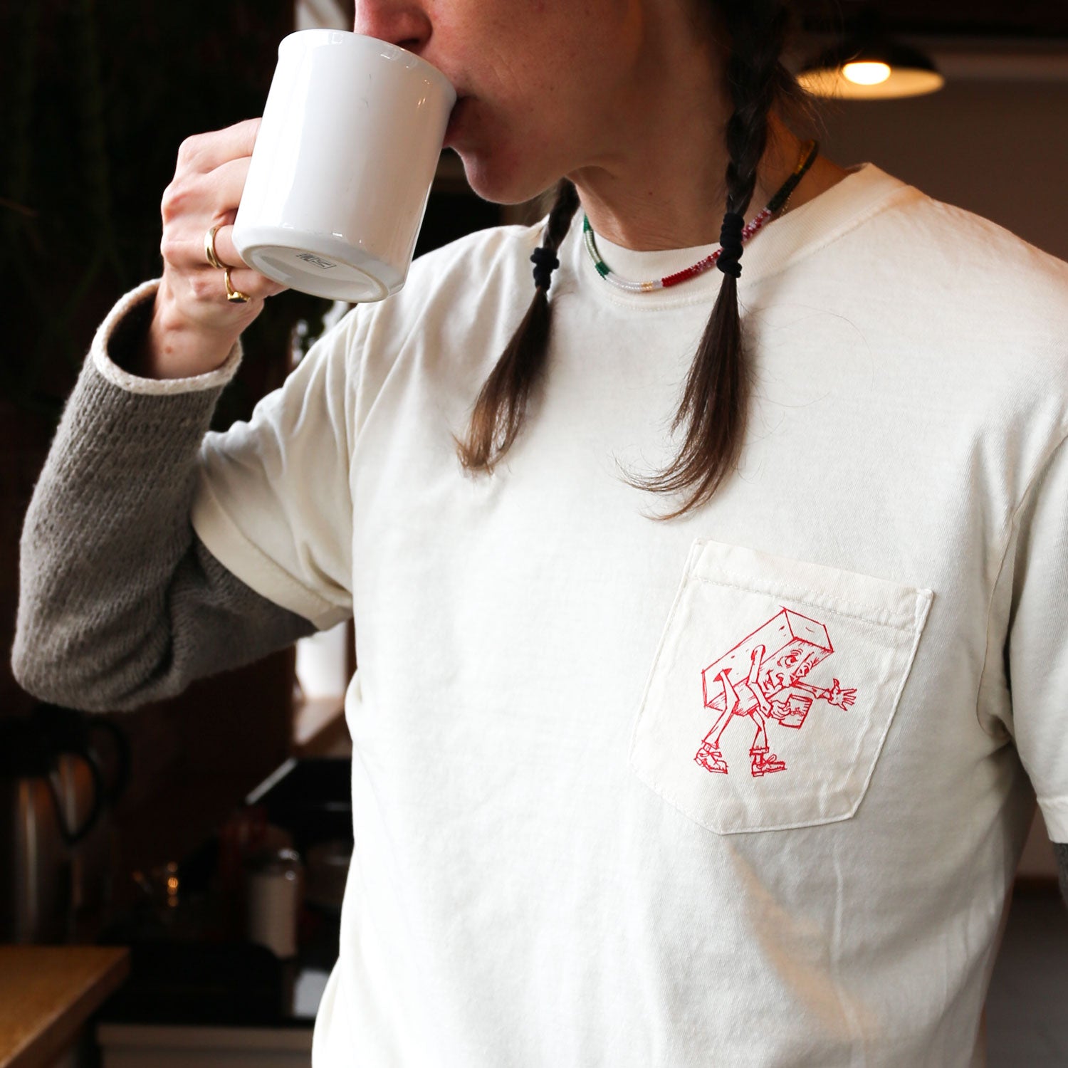 A person with braided hair, wearing the Brick Tee by Tandem Coffee Roasters featuring a red printed design on the pocket, is sipping from a white mug while flipping through Brick Journal. They are indoors, and part of their lower arm is covered by a gray sleeve.