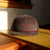 A Custom Tandem Coffee Roasters Corduroy Tandem Hat with a bicycle emblem embroidered on the front sits on a yellow table, bathed in warm sunlight with a shadow cast behind.