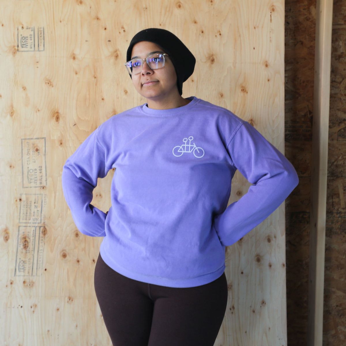 Woman in a Tandem Crewneck Sweatshirt and black beanie standing confidently with hands on hips in front of a wooden wall.