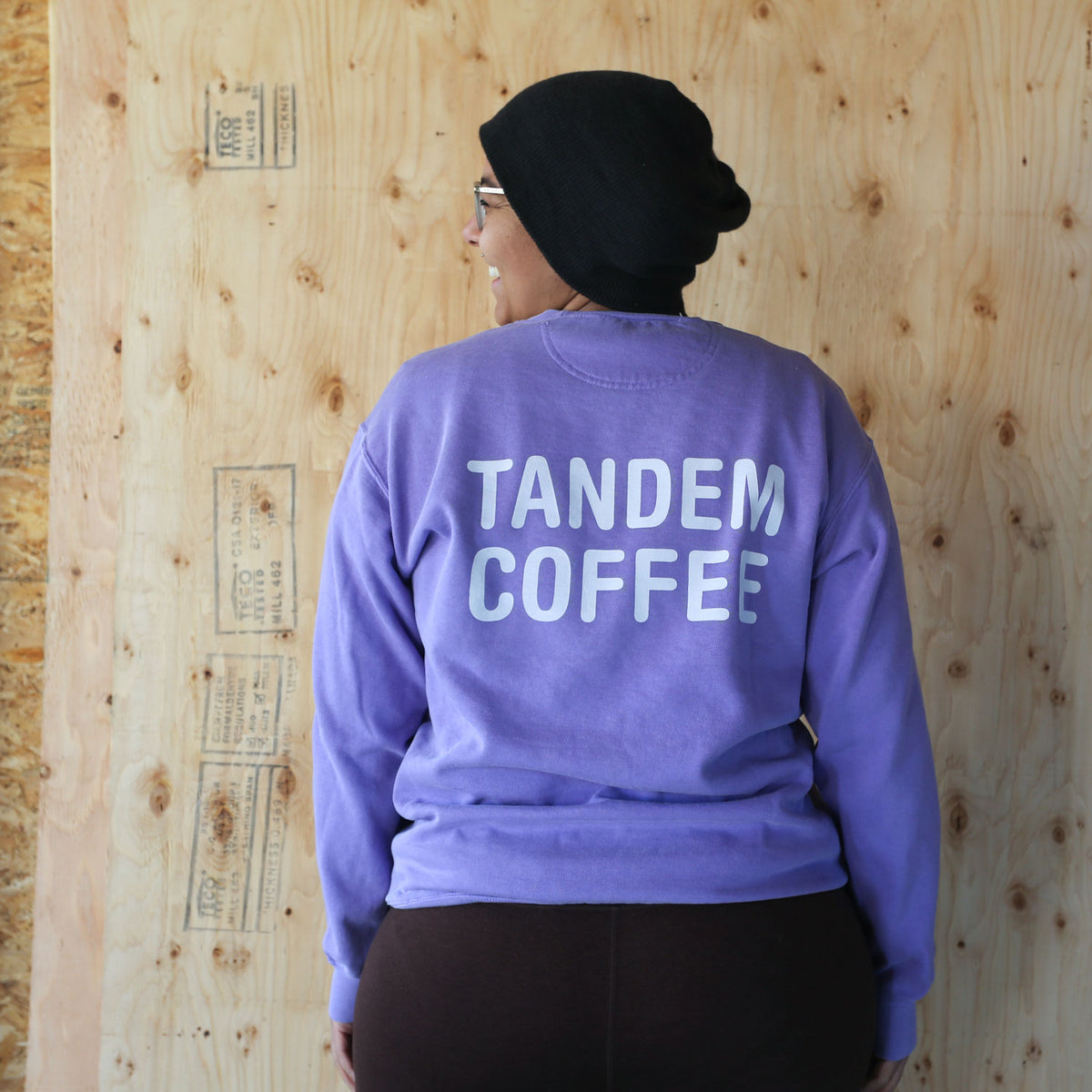 A person wearing a purple Tandem Coffee Roasters crewneck sweatshirt with "Tandem Coffee" printed on the back, facing a wooden plywood wall. The person is also wearing a black beanie and dark pants.