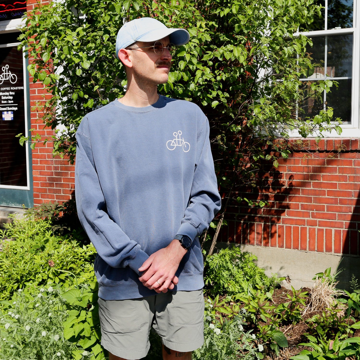 A person stands outdoors in front of a brick wall and green plants on a sunny day, wearing sunglasses and the blue Tandem Coffee Roasters Denim Tandem Crewneck sweatshirt along with a blue cap and gray shorts.