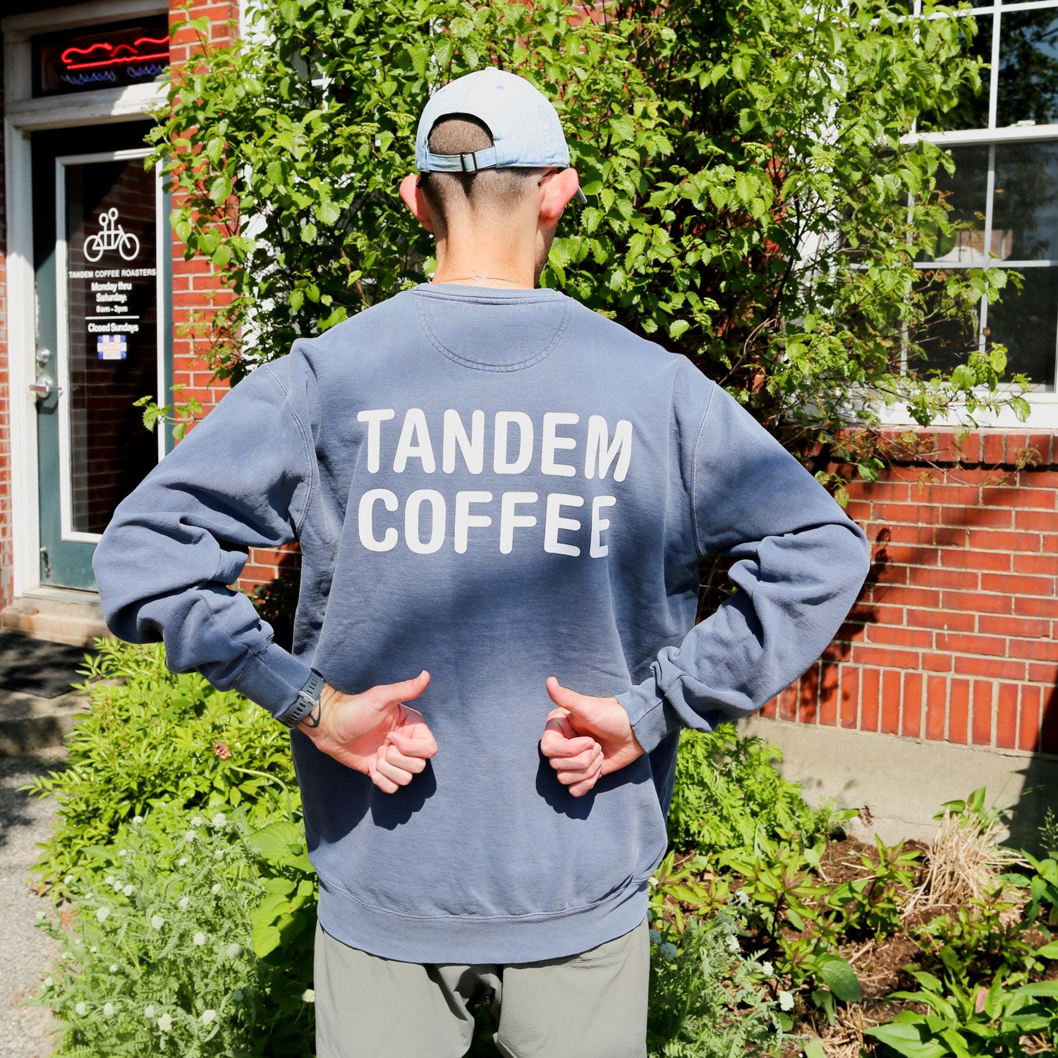 A person stands outdoors, back to the camera, giving two thumbs up while wearing a Denim Tandem Crewneck from Tandem Coffee Roasters. The sweatshirt features TANDEM COFFEE, with greenery and a brick building in the background.
