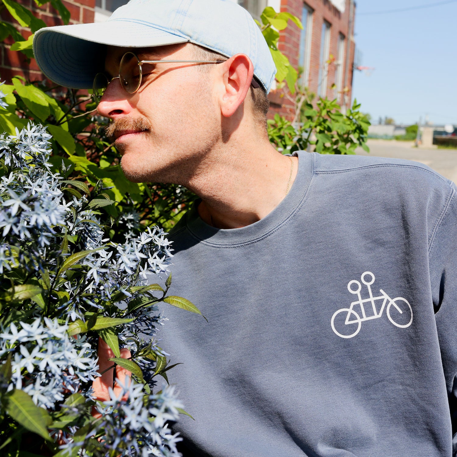 A man in a light blue cap and round glasses, wearing the Denim Tandem Crewneck by Tandem Coffee Roasters with a simple bicycle graphic, leans in to smell pale blue flowers near a brick building on a sunny day.