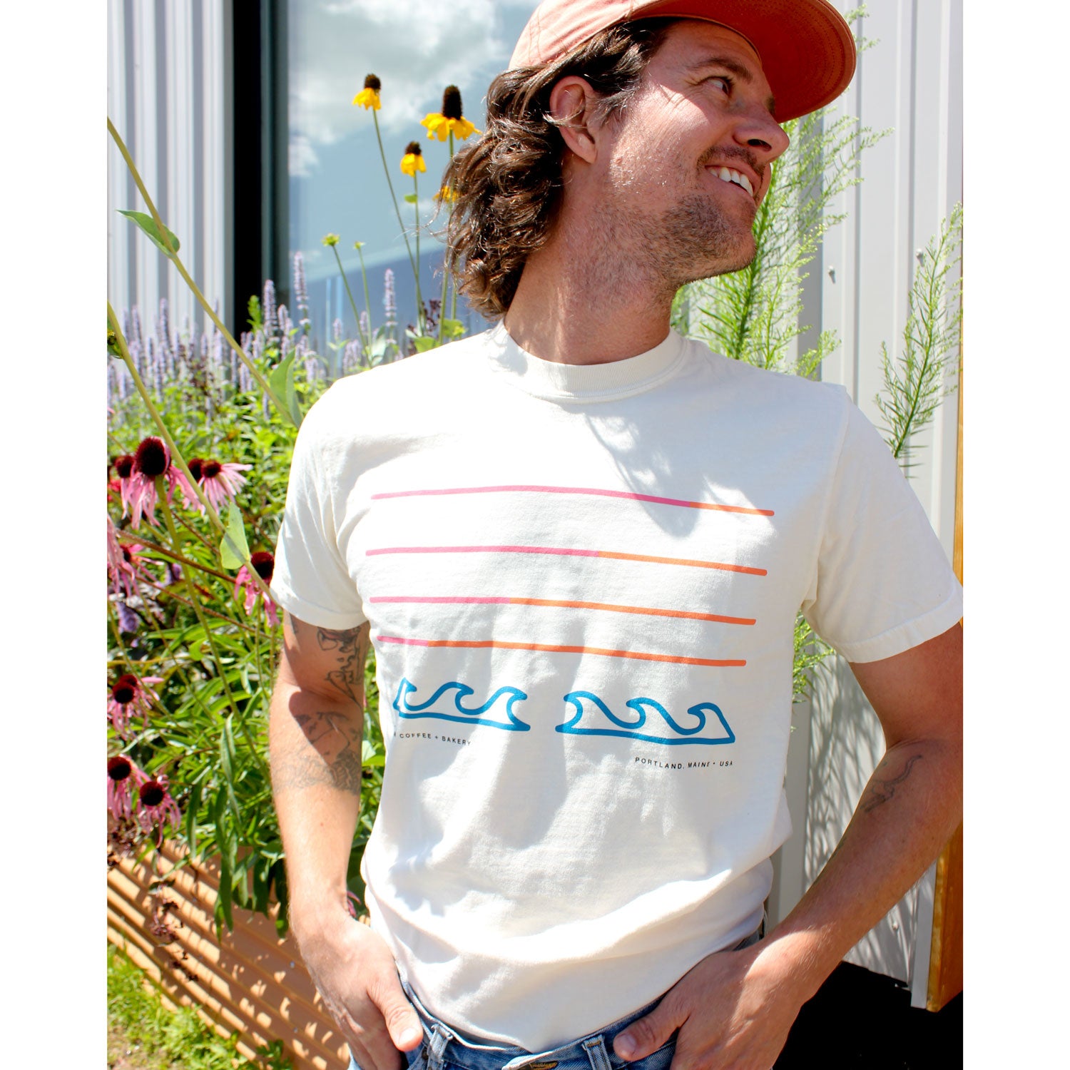 A smiling man in a cap and Tandems Filthy Sunset Tee stands outdoors by tall flowers and greenery, with a modern building and blue sky behind him. Limited stock—grab this unique Comfort Colors shirt while supplies last!.