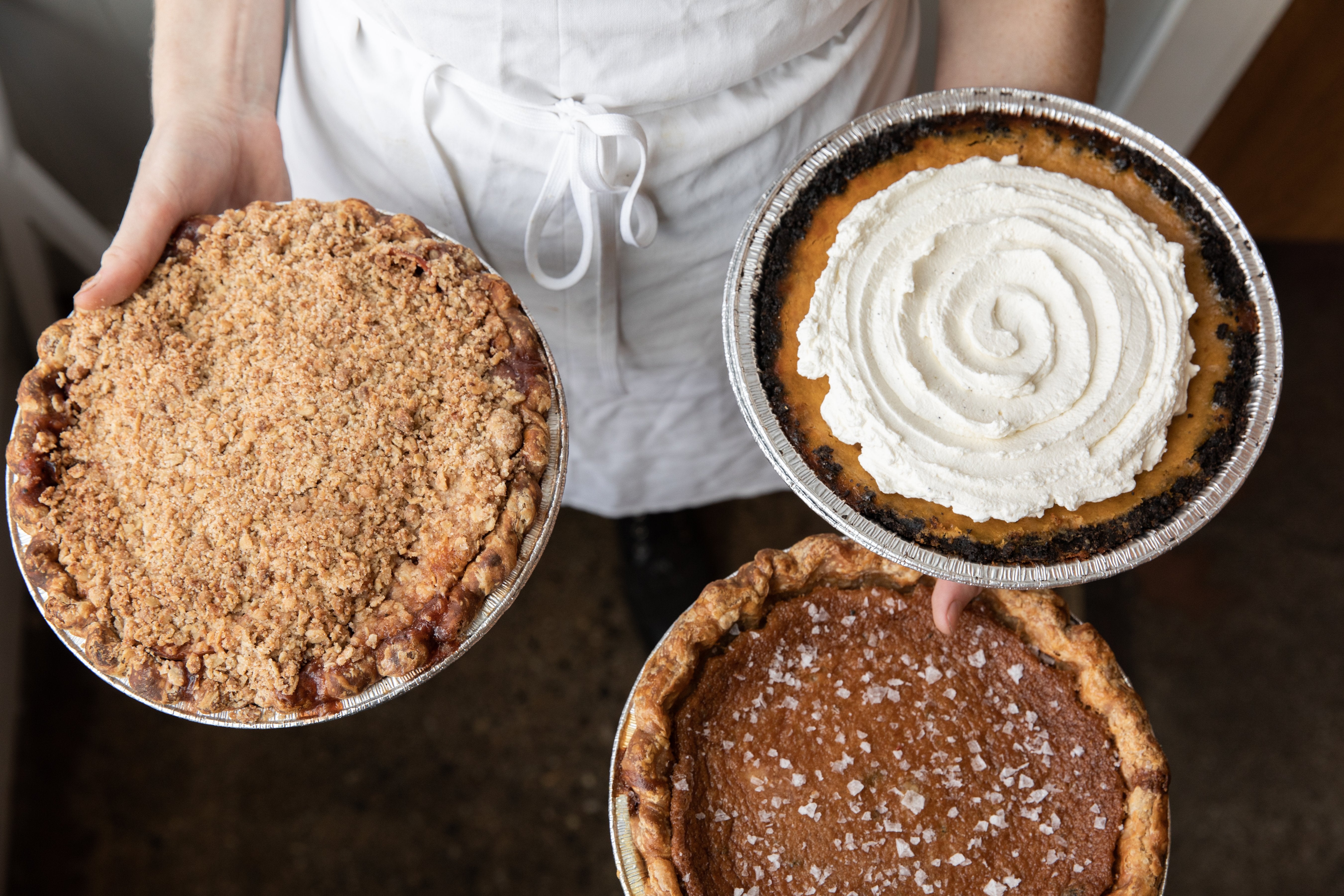 A person in a white apron holds three different pies: one with a crumble topping, another with whipped cream, and a third sprinkled with powdered sugar.