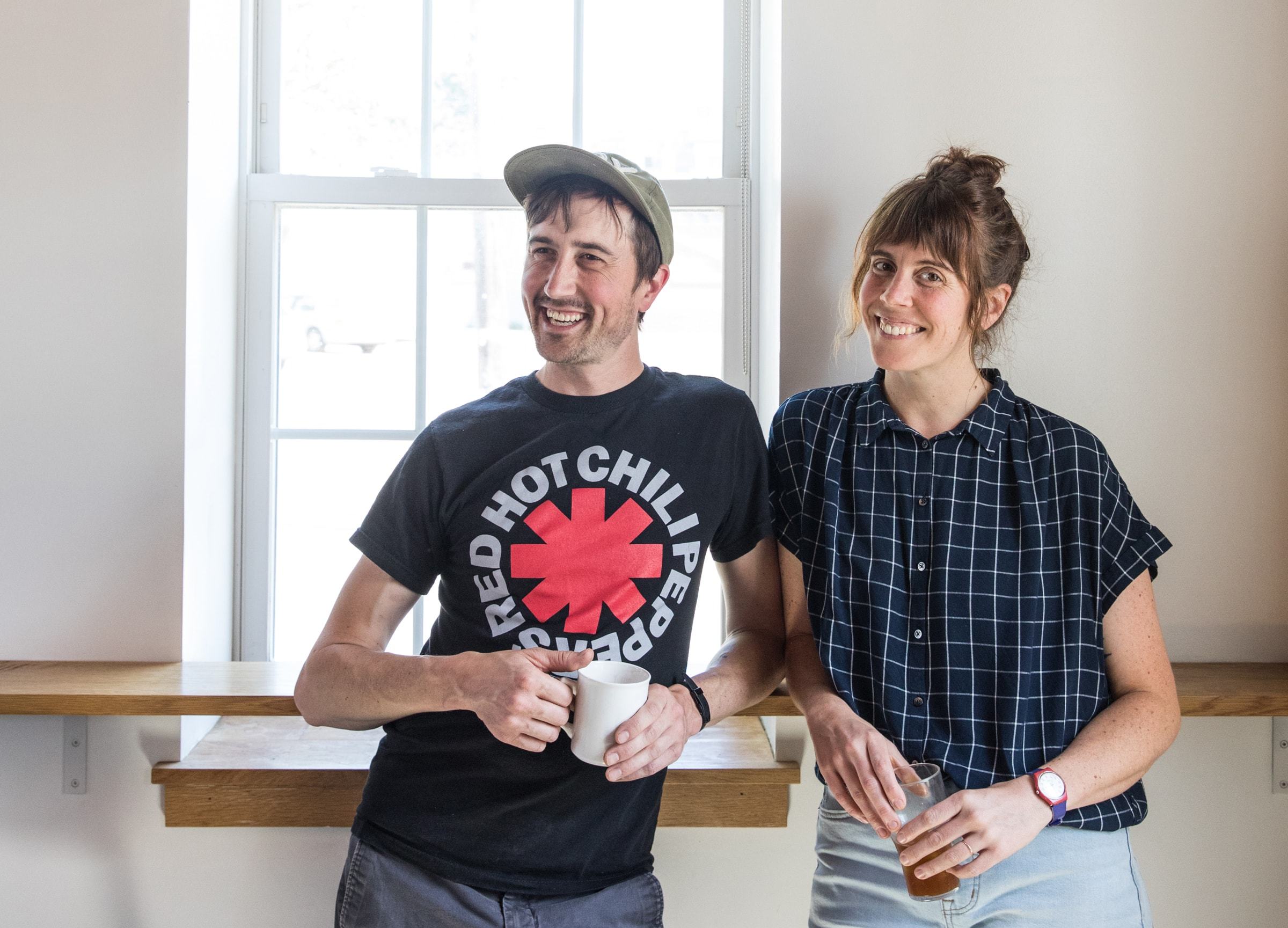 Two people are standing indoors in front of a window, smiling. The person on the left is wearing a Red Hot Chili Peppers t-shirt and cap while holding a white mug from Tandem Coffee. The person on the right is wearing a dark checkered shirt and holding a glass of what seems like iced tea, reminiscent of a relaxed afternoon in Portland, Maine.