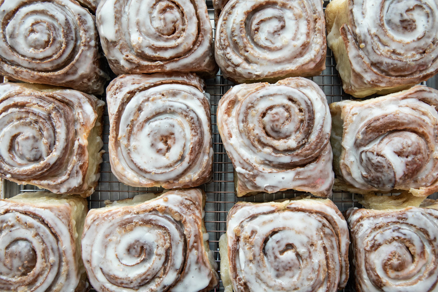 Freshly baked cinnamon rolls with a light layer of white icing are arranged in a grid pattern on a cooling rack at Tandem Coffee in Portland, Maine. The rolls are uniformly shaped with visible swirls of cinnamon filling. The icing glistens, giving the treats an appealing, mouth-watering appearance.