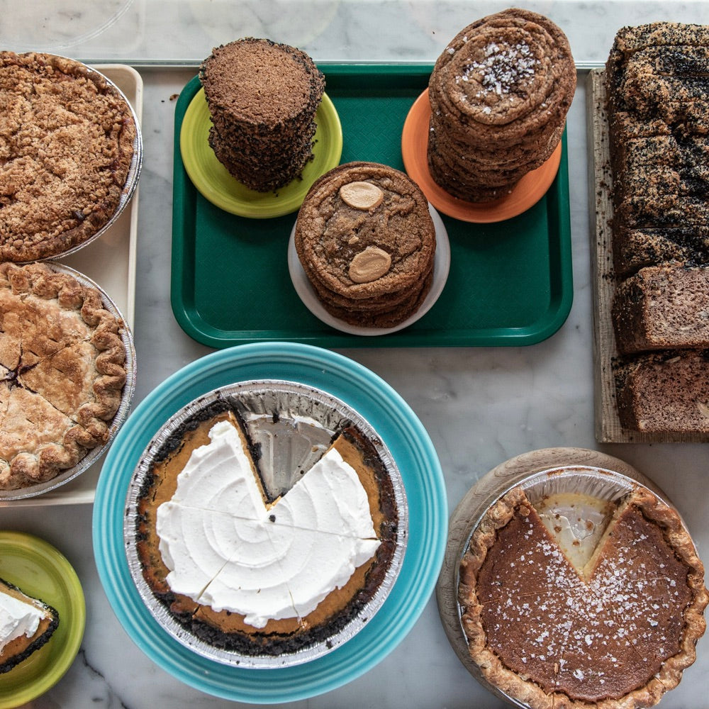 A variety of baked goods are displayed on a marble surface. There are pies with slices missing, stacks of cookies on colorful plates, a stack of brownies with a dusting of powdered sugar, and crumbly pastries. The assortment features different textures and colors, perfect for pairing with Tandem Coffee in Portland, Maine.
