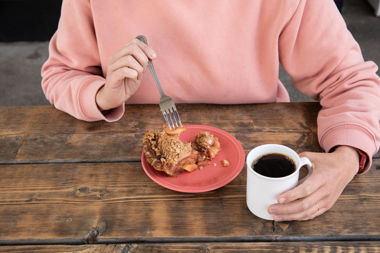 Person in a pink sweater holding a fork with a piece of dessert, possibly apple crumble, on a red plate. They are seated at a wooden table, holding a white mug filled with black coffee from Tandem Coffee in Portland, Maine, in their other hand.