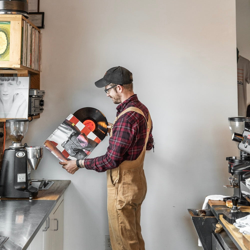 A man wearing a cap, glasses, and brown overalls over a red plaid shirt is holding a vinyl record in Tandem Coffee, Portland Maine. The record shelf behind him has several albums. Coffee brewing equipment and tools are on the counter beside him.