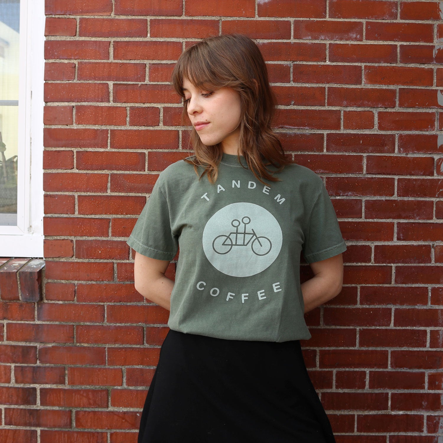 A person with shoulder-length brown hair leans against a brick wall, wearing a Moss Circle Logo Tee by Tandem Coffee Roasters, featuring the company's logo and a bicycle graphic. With hands behind their back, they gaze down to the side.