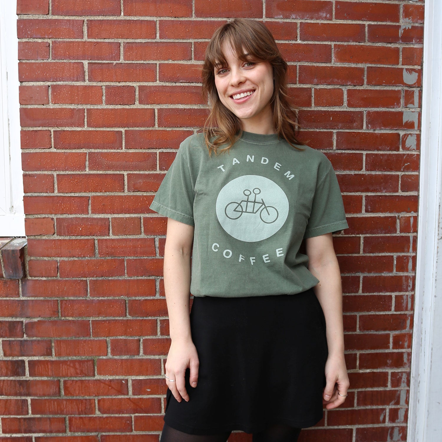 A person smiles in front of a brick wall, wearing the Moss Circle Logo Tee by Tandem Coffee Roasters, featuring a tandem bicycle graphic. They pair it perfectly with a black skirt.