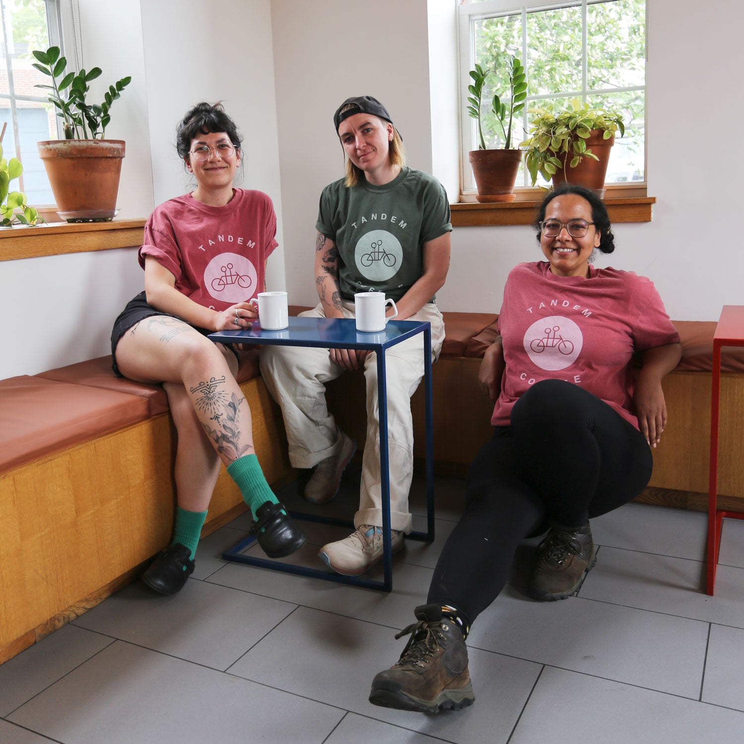 Three people are sitting at a table in a casual café setting, each wearing "Tandem Coffee Roasters" Crimson Circle Logo Tees in Dusty Crimson. They have mugs in front of them and are smiling at the camera. There are potted plants on a windowsill behind them.