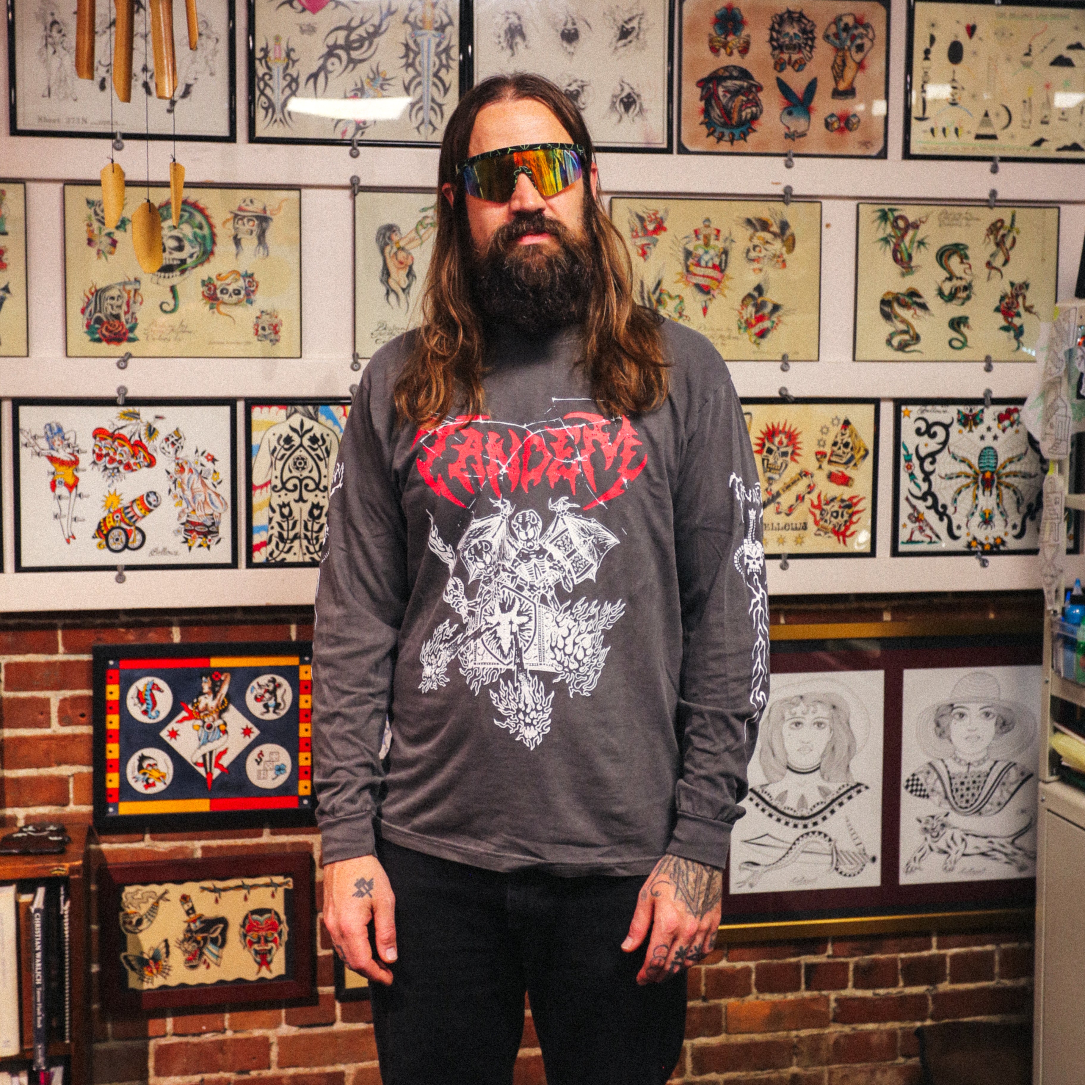 A bearded person in sunglasses and a Tandem Coffee Roasters Tandem Metal Long Sleeve stands in Gold Star Tattoo studio, with tattoo art covering the walls behind them.