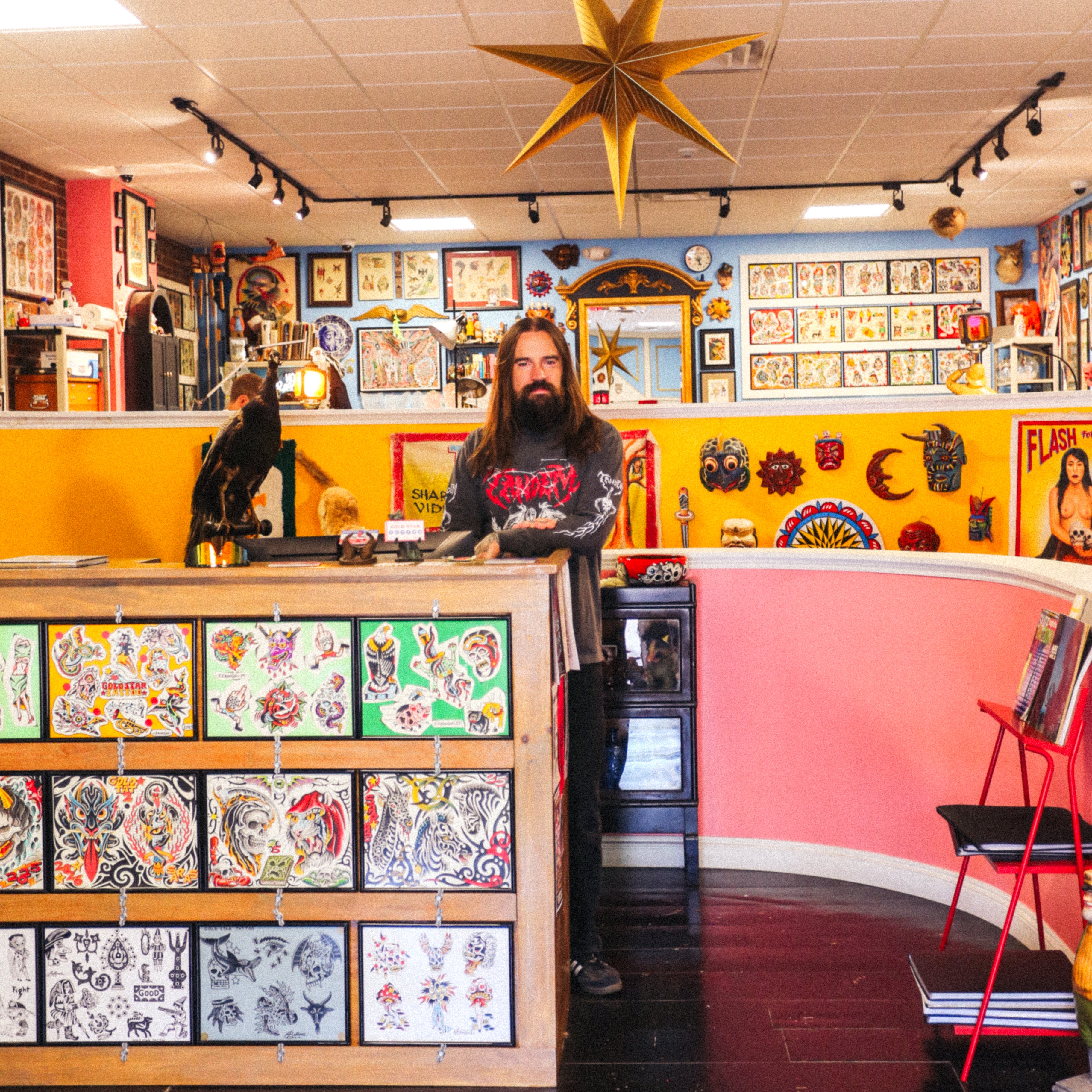 A long-haired person stands behind the Gold Star Tattoo shop counter, surrounded by tattoo flash art, vibrant walls, and a gold star hanging from the ceiling. Tattoo designs are displayed on the counter and along the wall, while the artist wears a Tandem Metal Long Sleeve from Tandem Coffee Roasters that perfectly complements the creative atmosphere.