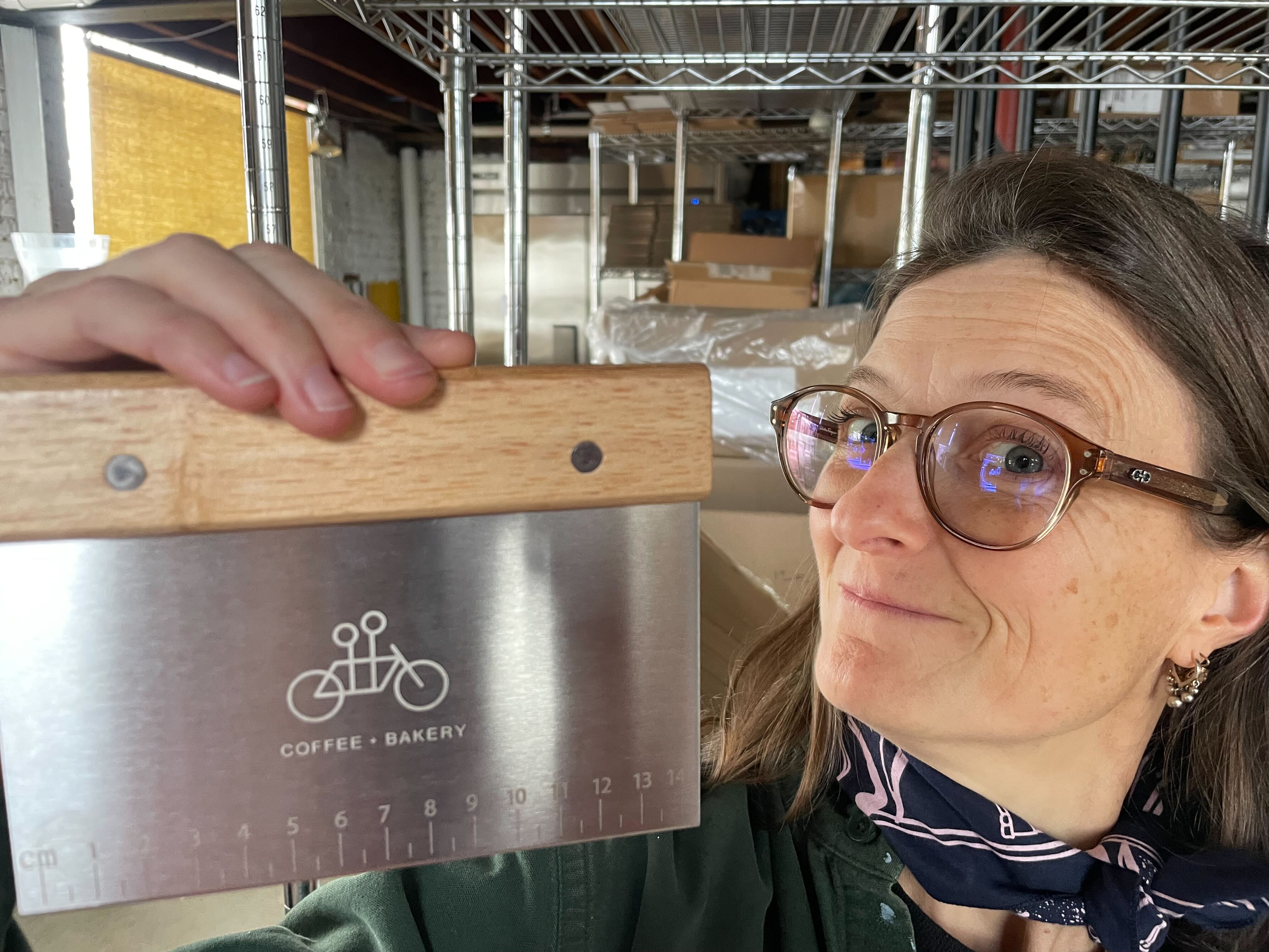 A woman in glasses holds up the Tandem Bench Scraper by Tandem Coffee Roasters, which has a wooden handle and bicycle logo, standing in a warehouse with shelves and cardboard boxes.