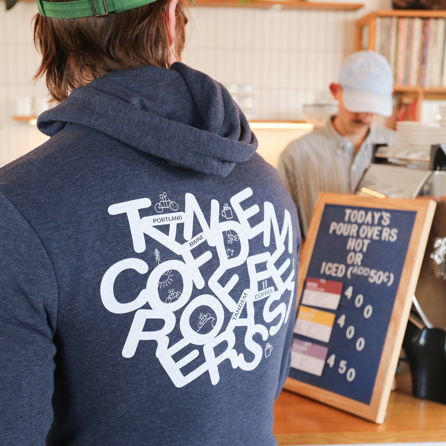 A person with long hair and a backward cap wears a stylish navy blue Letter Zip-Up Hoodie featuring "Tandem Coffee Roasters" on the back. They stand at a counter, where today's pour-over options are highlighted on the menu. In the background, another person diligently operates a coffee machine.