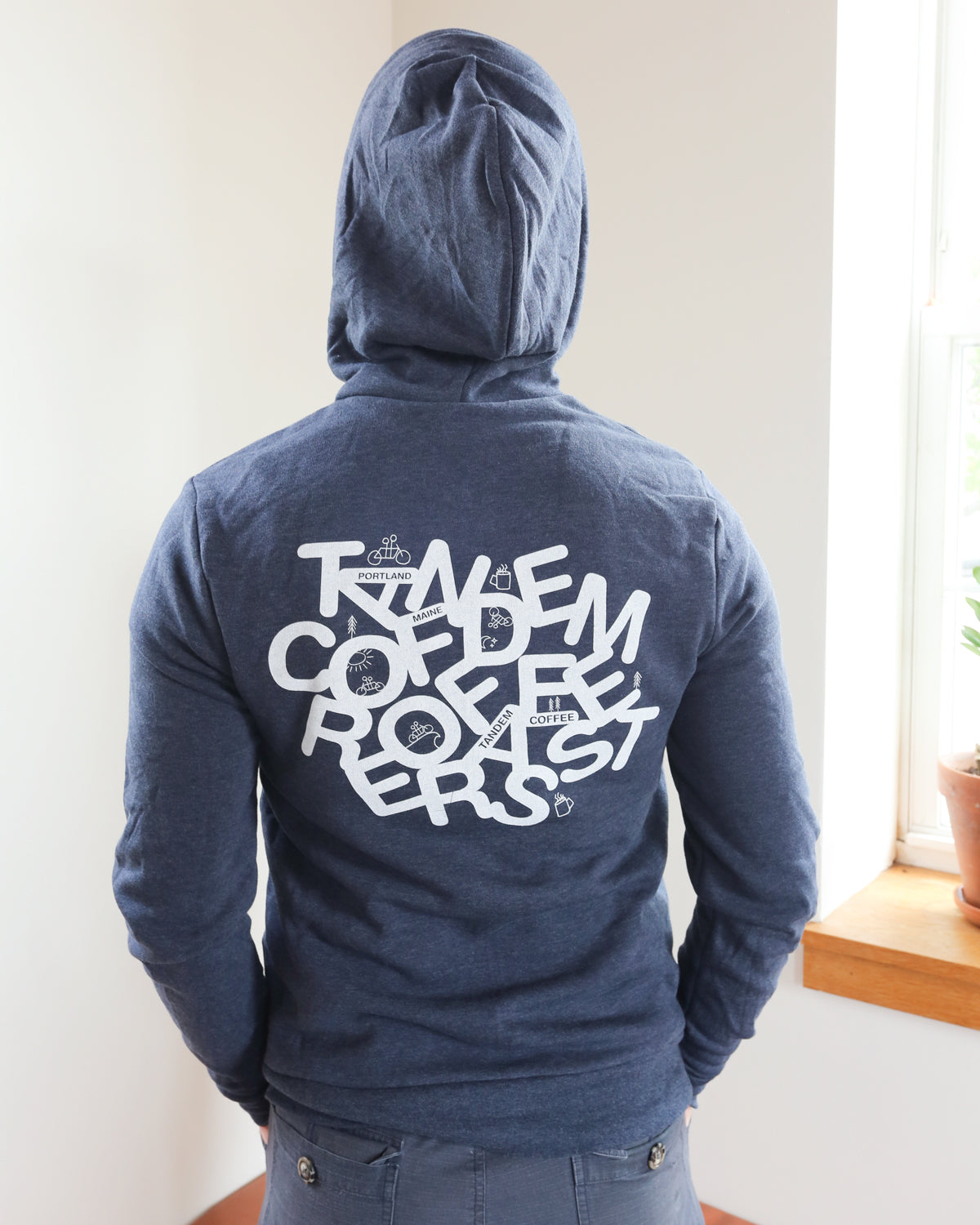 A person wearing a Tandem Coffee Roasters Letter Zip-Up Hoodie in navy blue stands facing a window. The stylish hoodie features white, stylized text with overlapping letters on the back, which are difficult to read. A small plant can be seen on the windowsill.