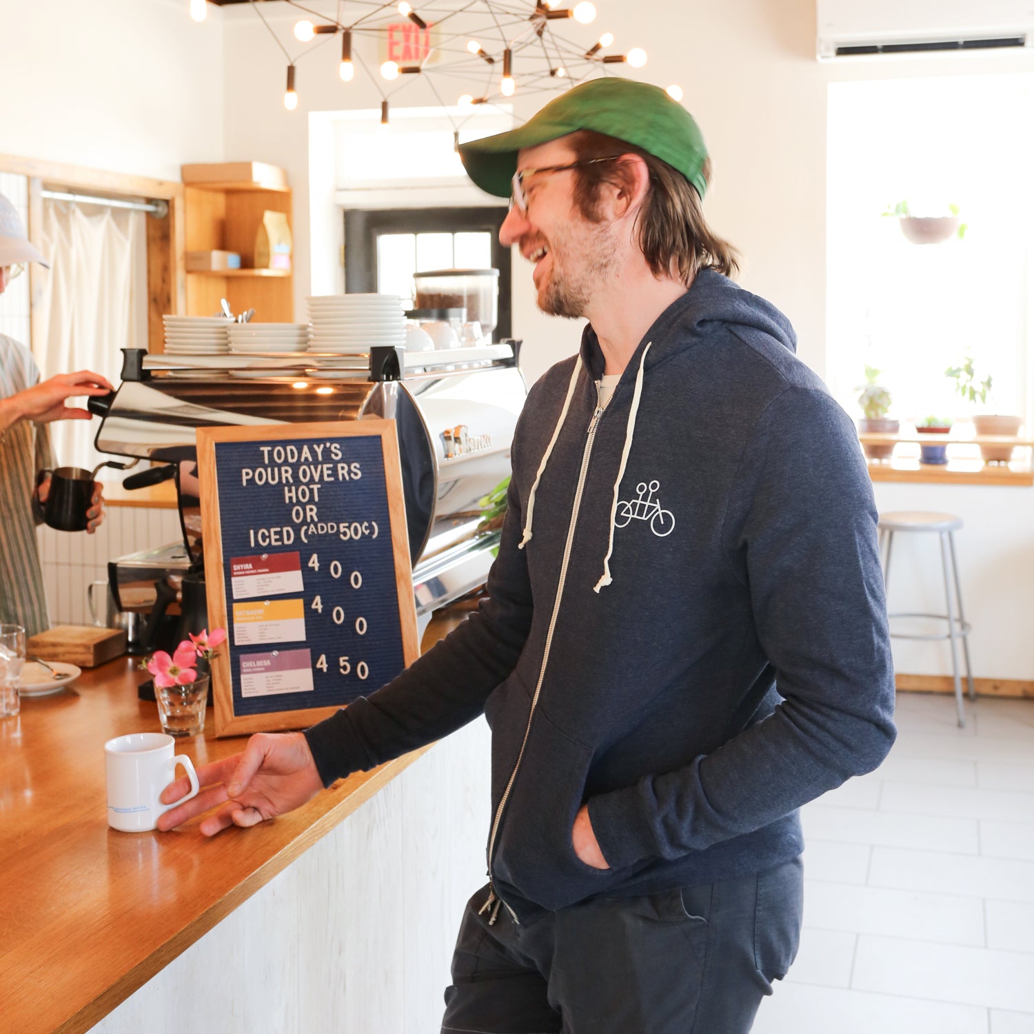 Dressed in a chic navy blue "Letter Zip-Up Hoodie" by Tandem Coffee Roasters, a man with a green cap stands in a cozy cafe, holding a mug. Beside him is the menu board for "Today's Pour Overs," displaying prices for hot and iced coffee options. Behind him, a barista busily crafts drinks.