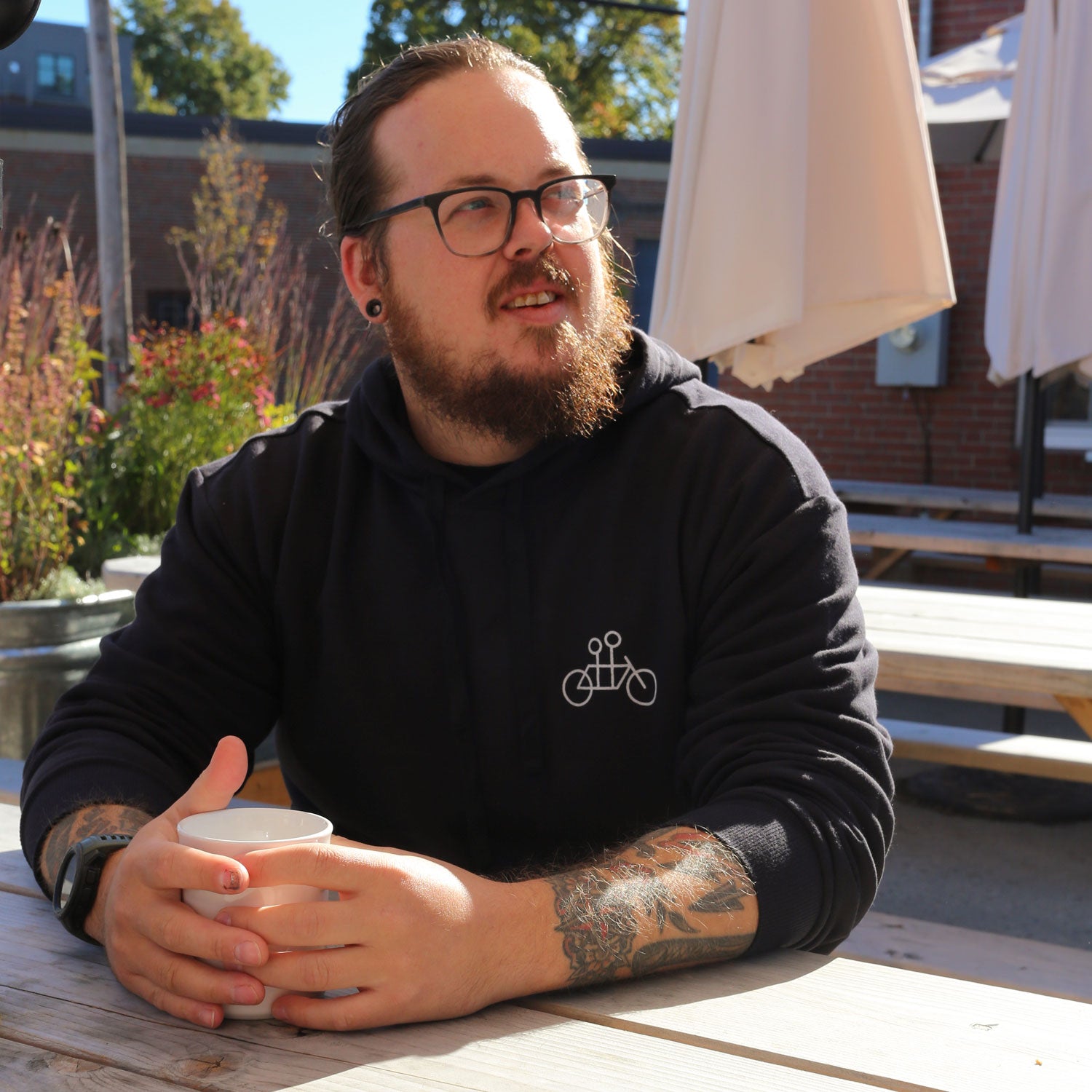 A person with glasses and tattoos sits at a picnic table outdoors, holding a cup. They wear a Tandem Coffee Roasters Letter Pullover Hoodie under an umbrella, with sunlight filtering through. A brick building and plants are in the background.