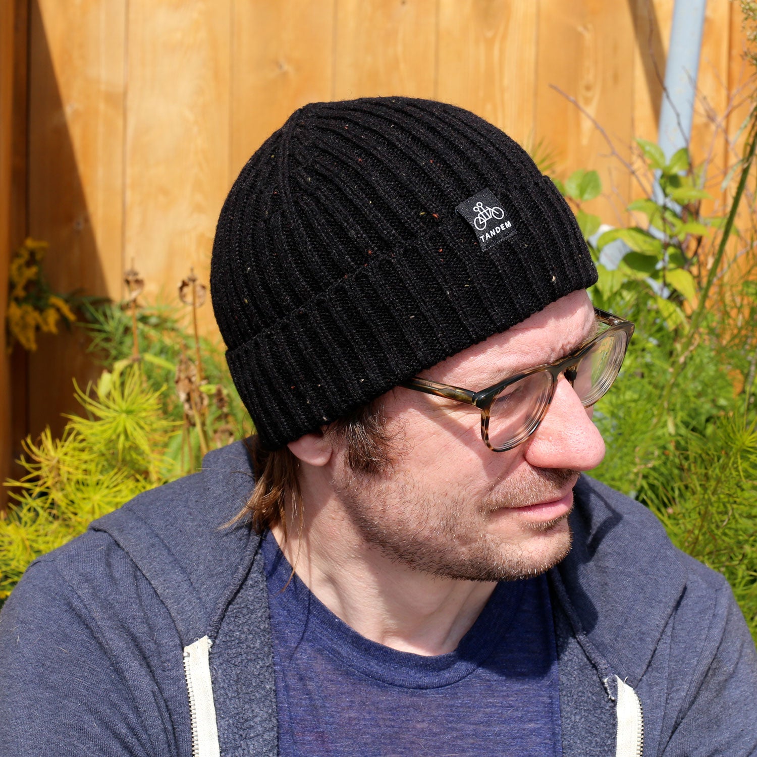A man wearing the Tandem Coffee Roasters Merino Wool Beanie, eyeglasses, and a blue hoodie sits outdoors near greenery and a wooden fence, looking to the side in natural sunlight.