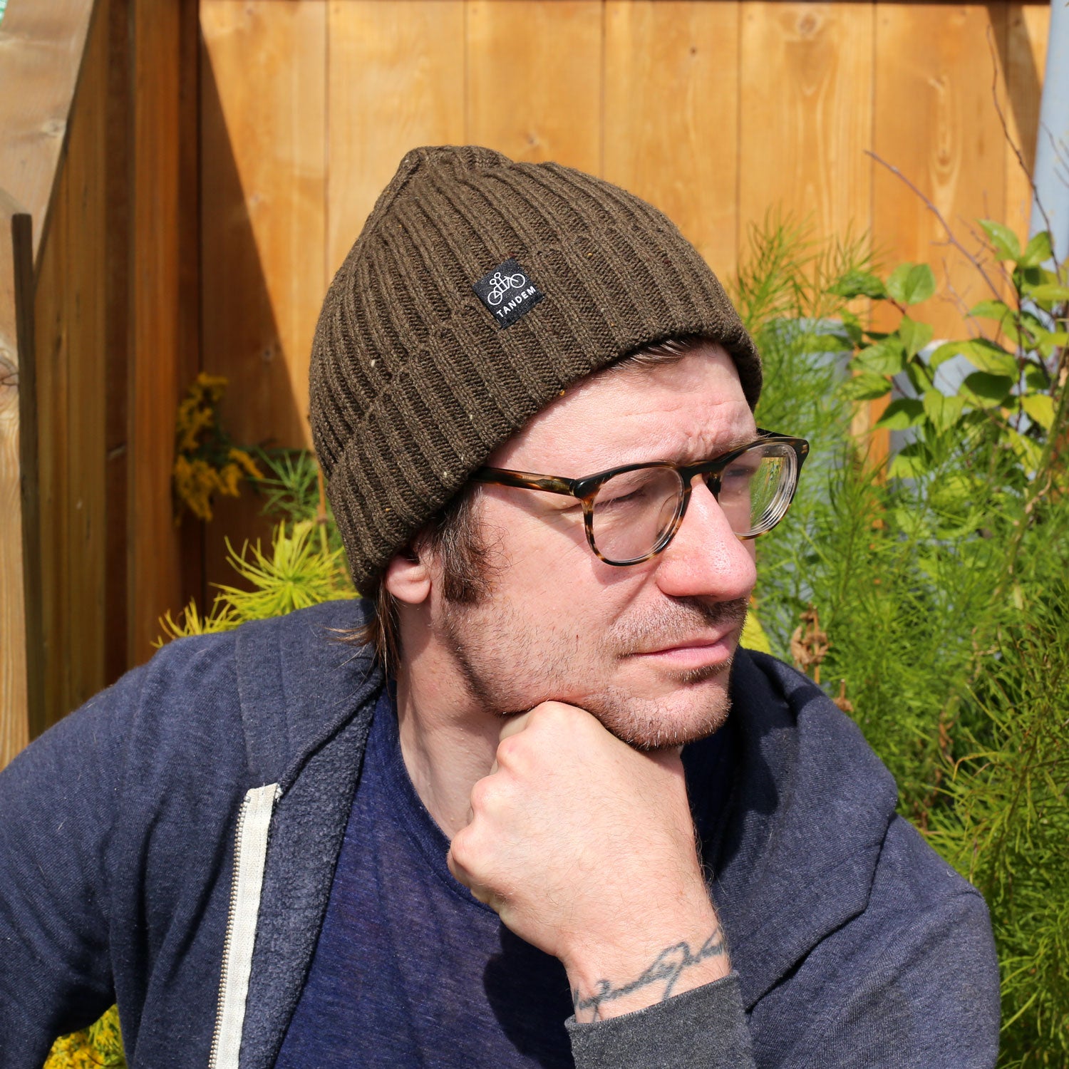 A man in glasses and a brown Merino Wool Beanie from Tandem Coffee Roasters, along with a blue hoodie, sits outdoors in sunlight, resting his chin on his hand thoughtfully. Green plants and a wooden fence are behind him.