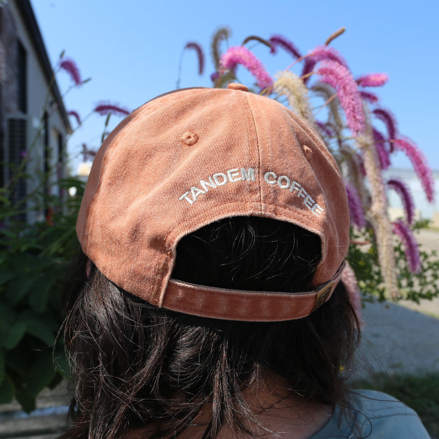 A person with long dark hair wears a Rust Circle Logo Dad Hat from Tandem Coffee Roasters, standing outdoors by tall pink flowers and greenery under a clear blue sky.
