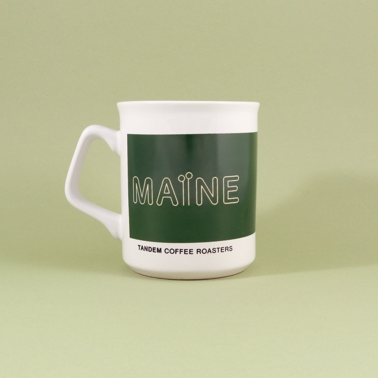 An 11oz white ceramic Maine Mug with a green band featuring the word "maine" in white, and "Tandem Coffee Roasters" written underneath, standing against a light green