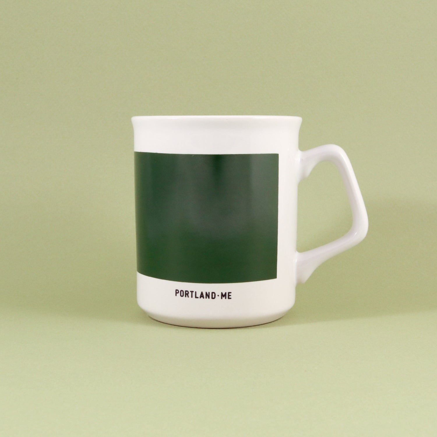 An 11oz white ceramic Maine Mug with a green band featuring the word "maine" in white, and "Tandem Coffee Roasters" written underneath, standing against a light green