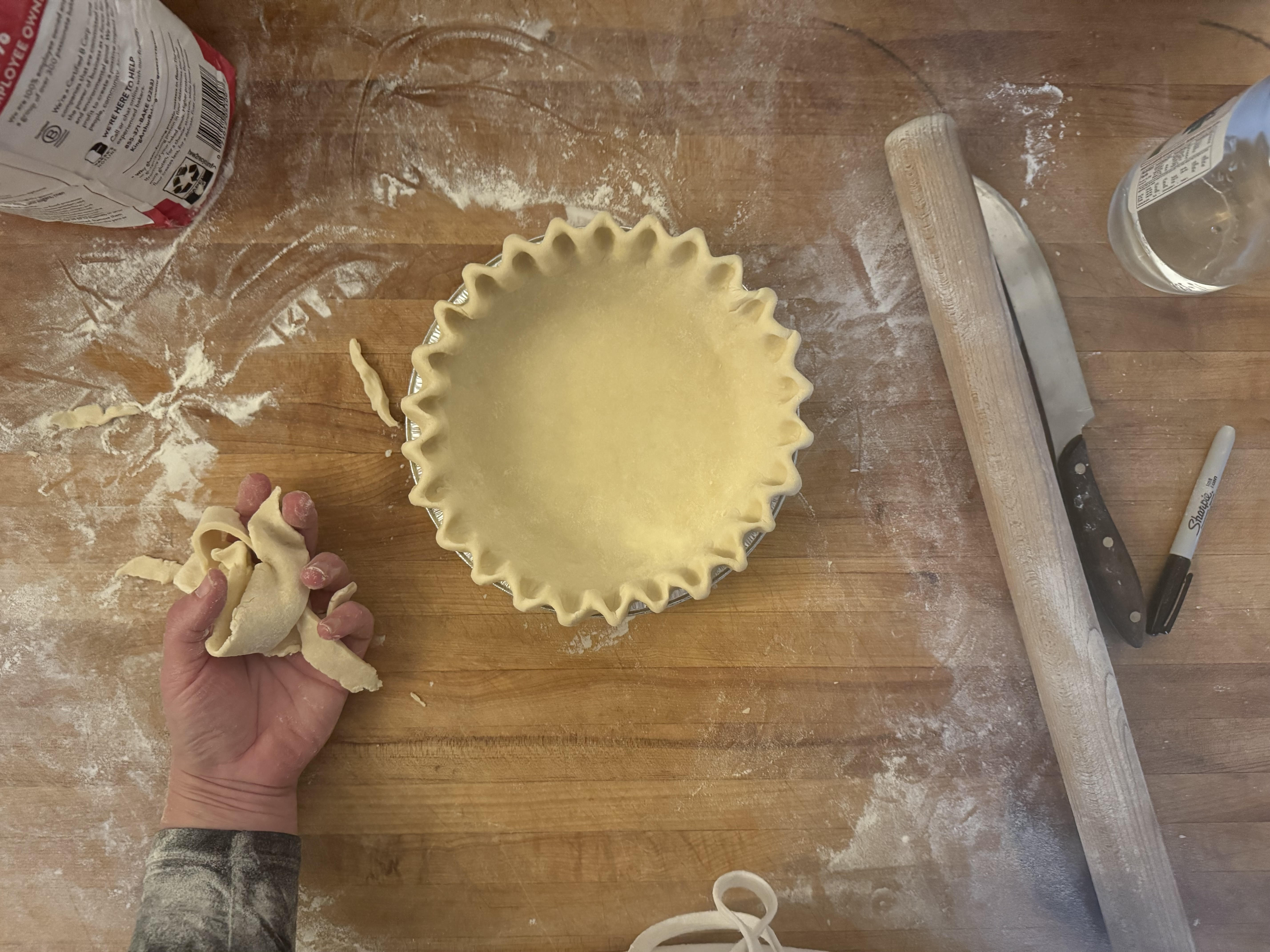 Person making pie crust on a wooden table with rolling pin and knife.