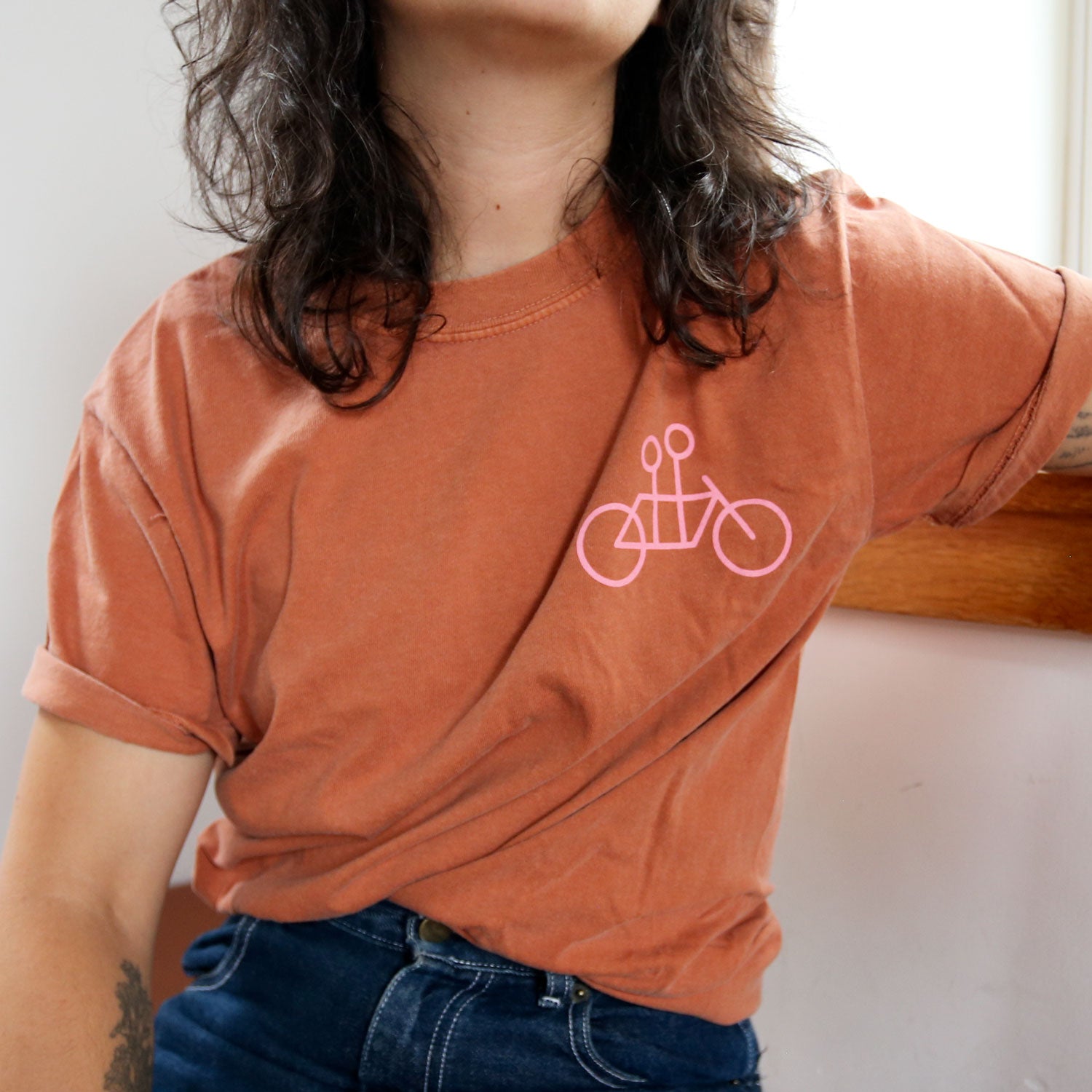 A person with long, wavy hair is wearing a rust-colored Tandem Coffee Roasters tee featuring a minimalist pink bicycle design on the chest. They are leaning against a wooden surface, with one arm resting on it. The photo is cropped at the shoulders and waist.