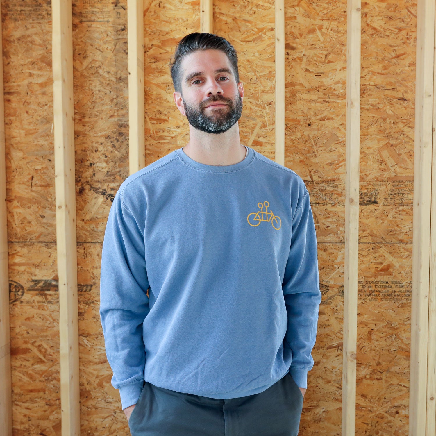 A man with a beard stands smiling in a blue Tandem Coffee Roasters crewneck sweatshirt with a Tandem Coffee bicycle logo, in front of a plywood wall in a construction site setting.