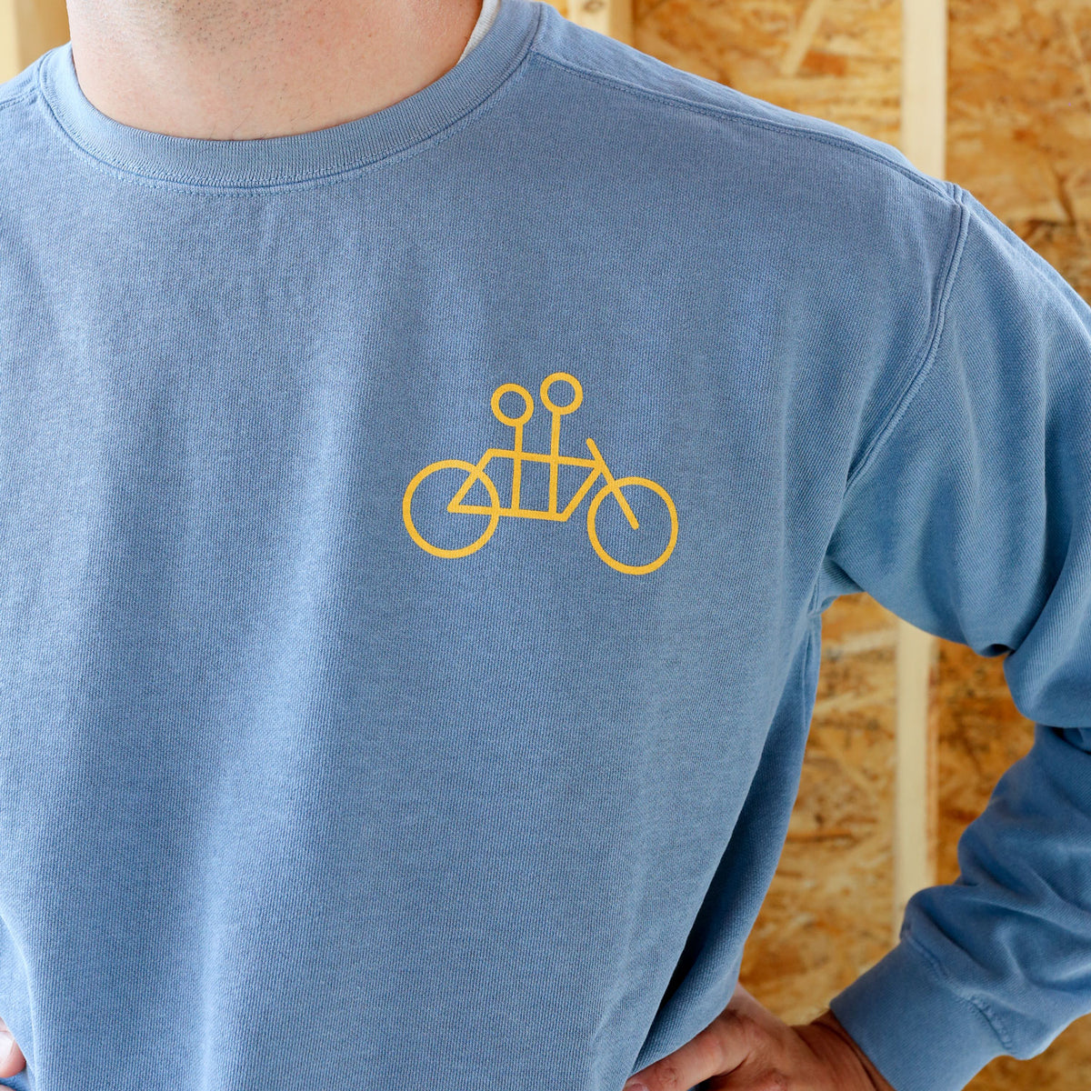 Close-up of a person wearing a Tandem Crewneck Sweatshirt by Tandem Coffee Roasters featuring a yellow minimalist bicycle design on the chest, standing against a wooden background.