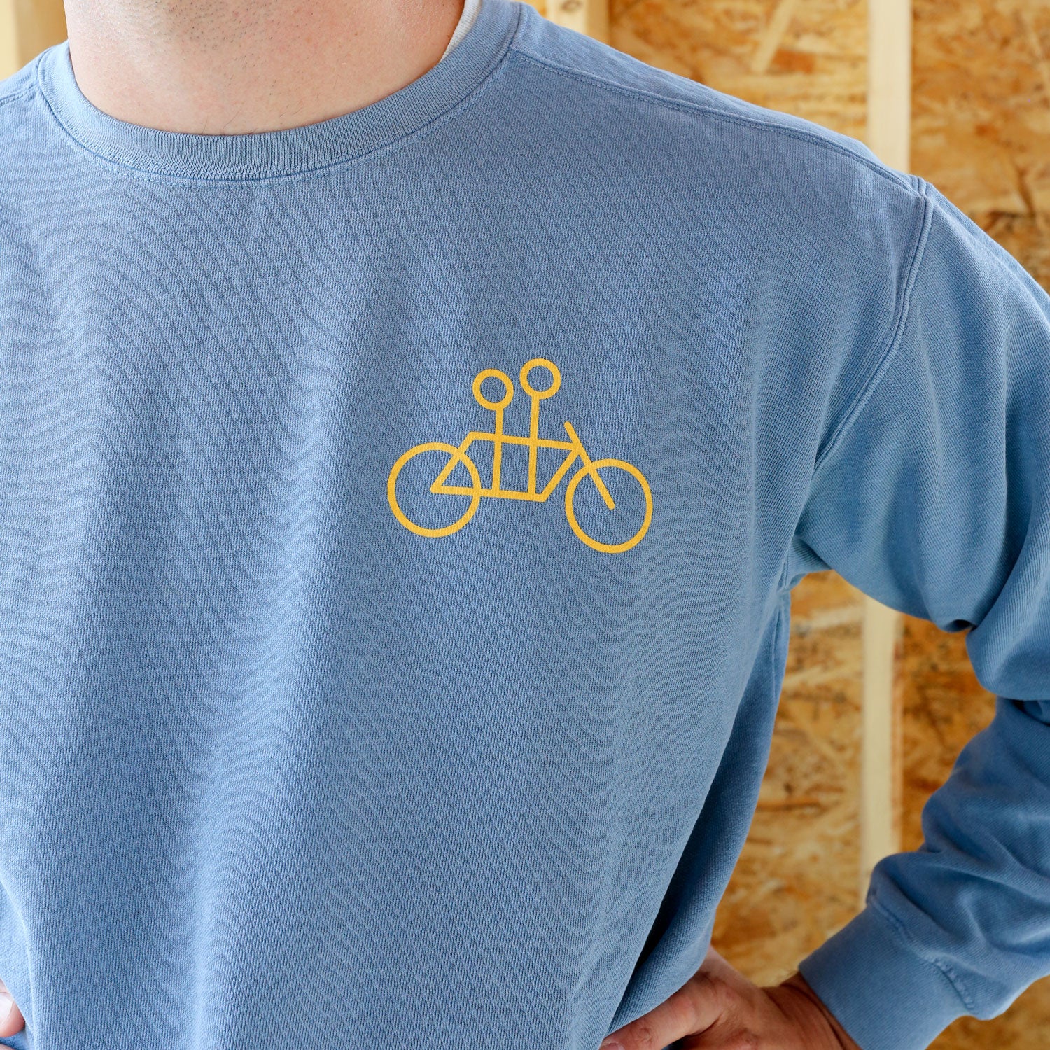 A man with a beard stands smiling in a blue Tandem Coffee Roasters crewneck sweatshirt with a Tandem Coffee bicycle logo, in front of a plywood wall in a construction site setting.