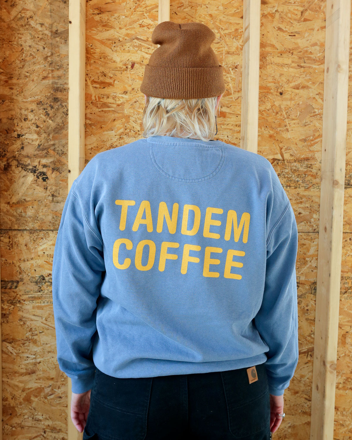 A person seen from behind wearing a blue Tandem Coffee Roasters crewneck sweatshirt with "Tandem Coffee" printed in yellow letters, a brown beanie, and black pants, standing in front of a plywood wall.
