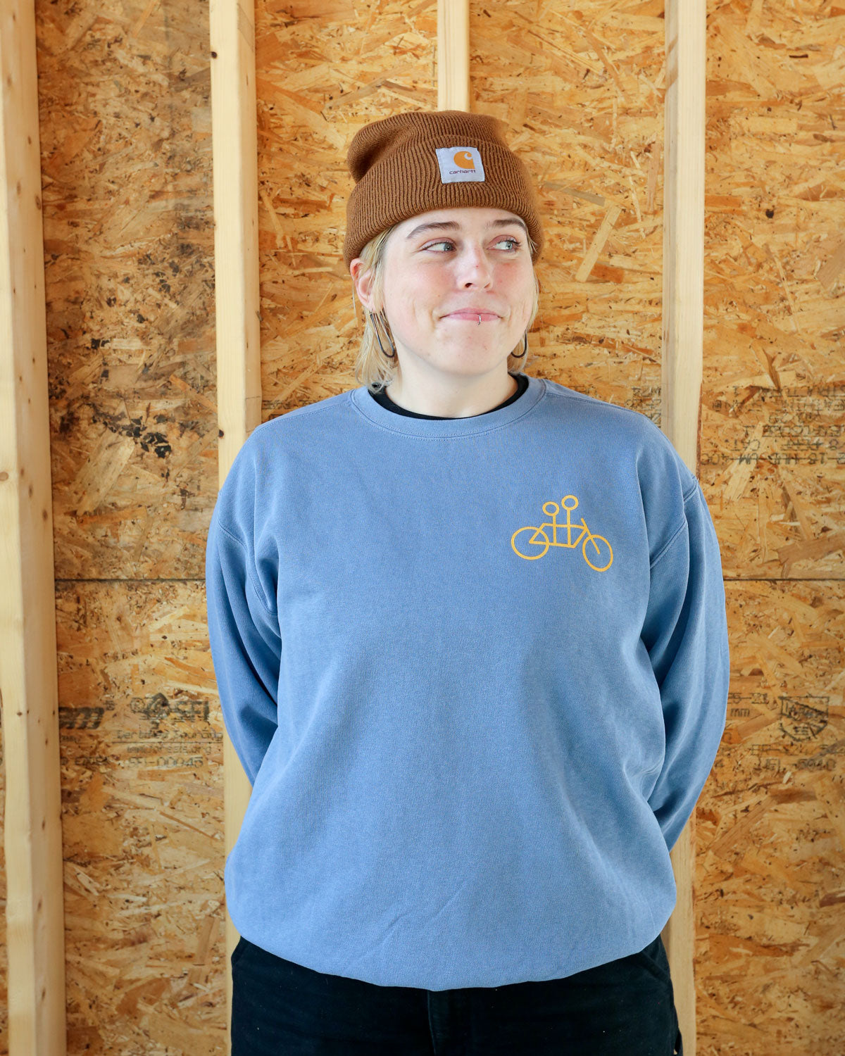 A person wearing a Tandem Crewneck Sweatshirt with a bicycle graphic and a brown beanie stands smiling against a plywood wall background.