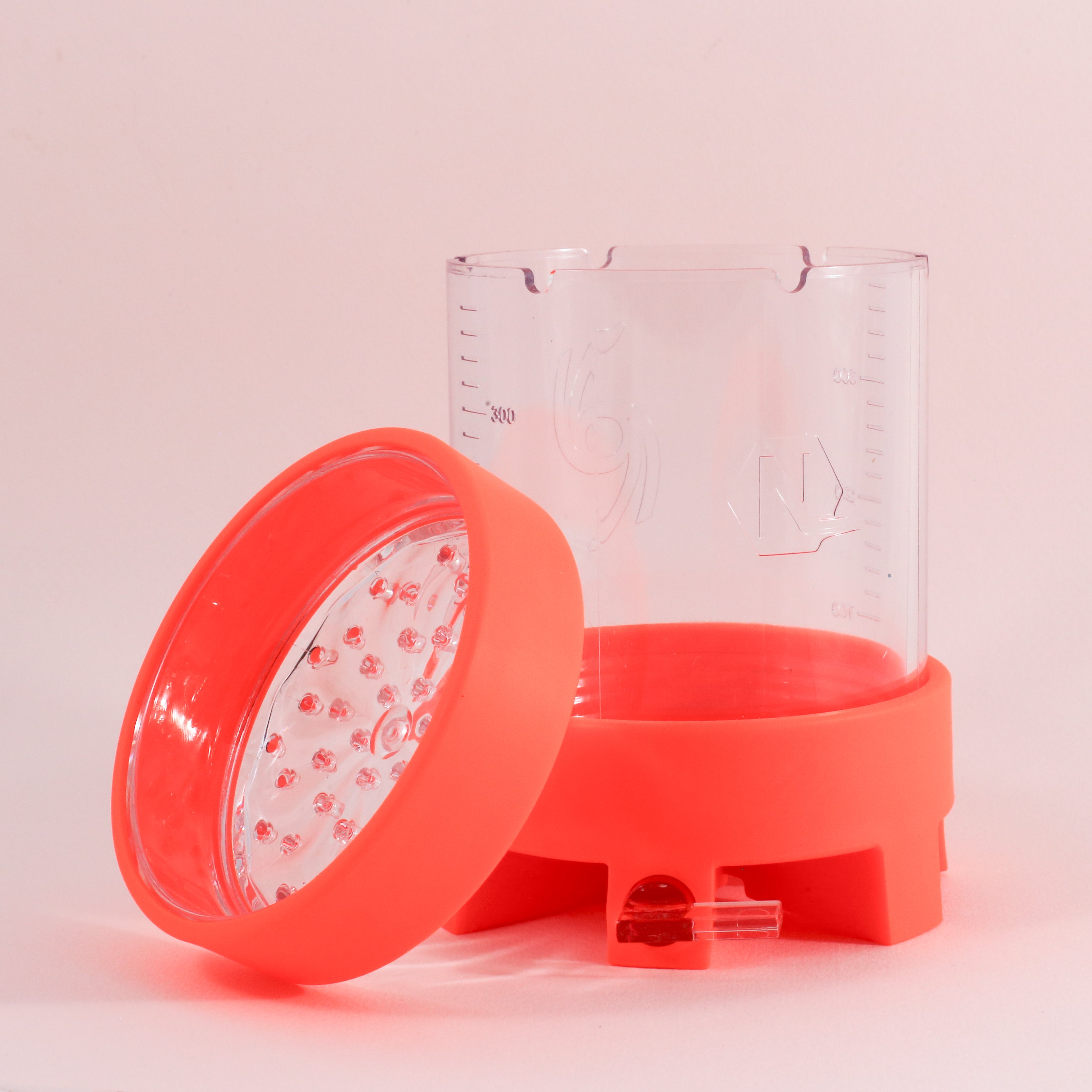 The NextLevel Pulsar Brewer by vendor-unknown is a clear plastic manual coffee maker with measurement markings, a bright orange base and lid with small holes, and an adjustable flow valve for precise brewing, shown on a light pink background.