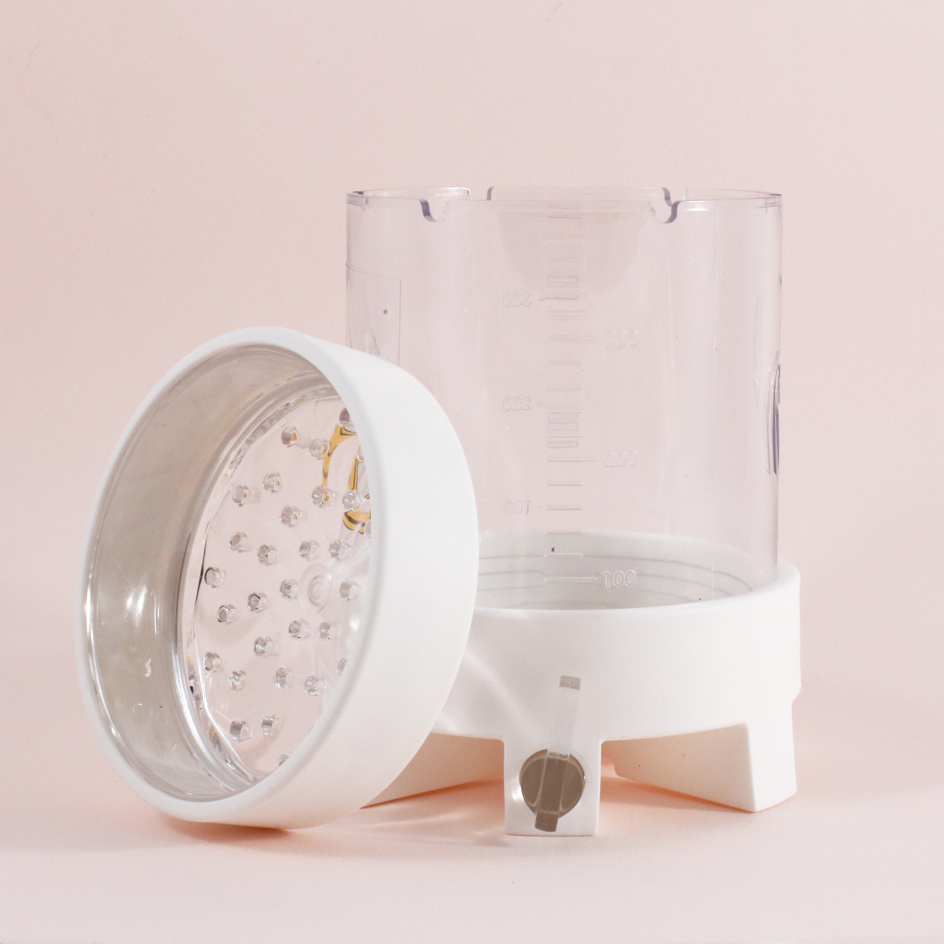 The NextLevel Pulsar Brewer by vendor-unknown features a clear plastic container with measurement markings and a detachable white lid with a metallic interior, small holes, and an adjustable flow valve, shown on a light pink background.