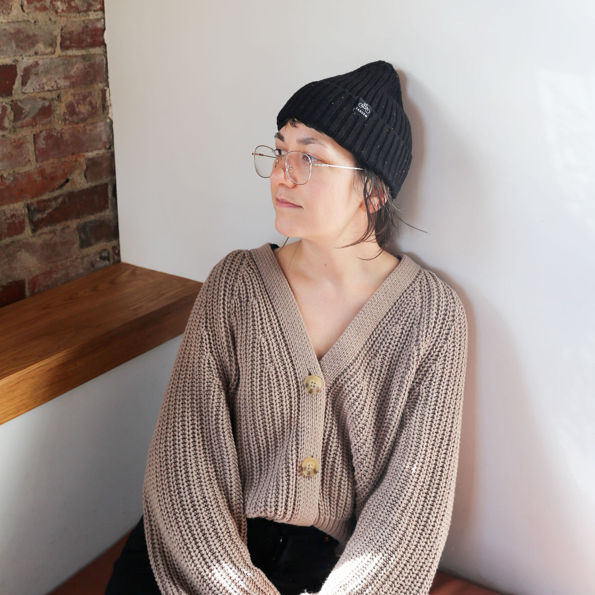 A woman wearing a black Tandem Coffee Roasters merino wool beanie and glasses sits at a wooden table against a brick wall background, looking to her right with a thoughtful expression. She's dressed in a chunky knit.
