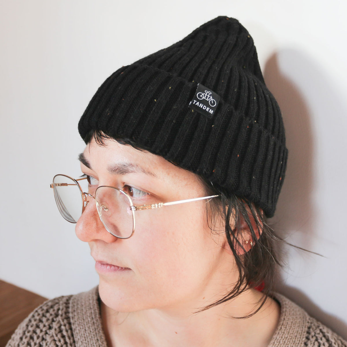 A person with short hair wearing glasses and a black ribbed Tandem Coffee Roasters merino wool beanie with a logo on the front, looking to the side against a plain background.